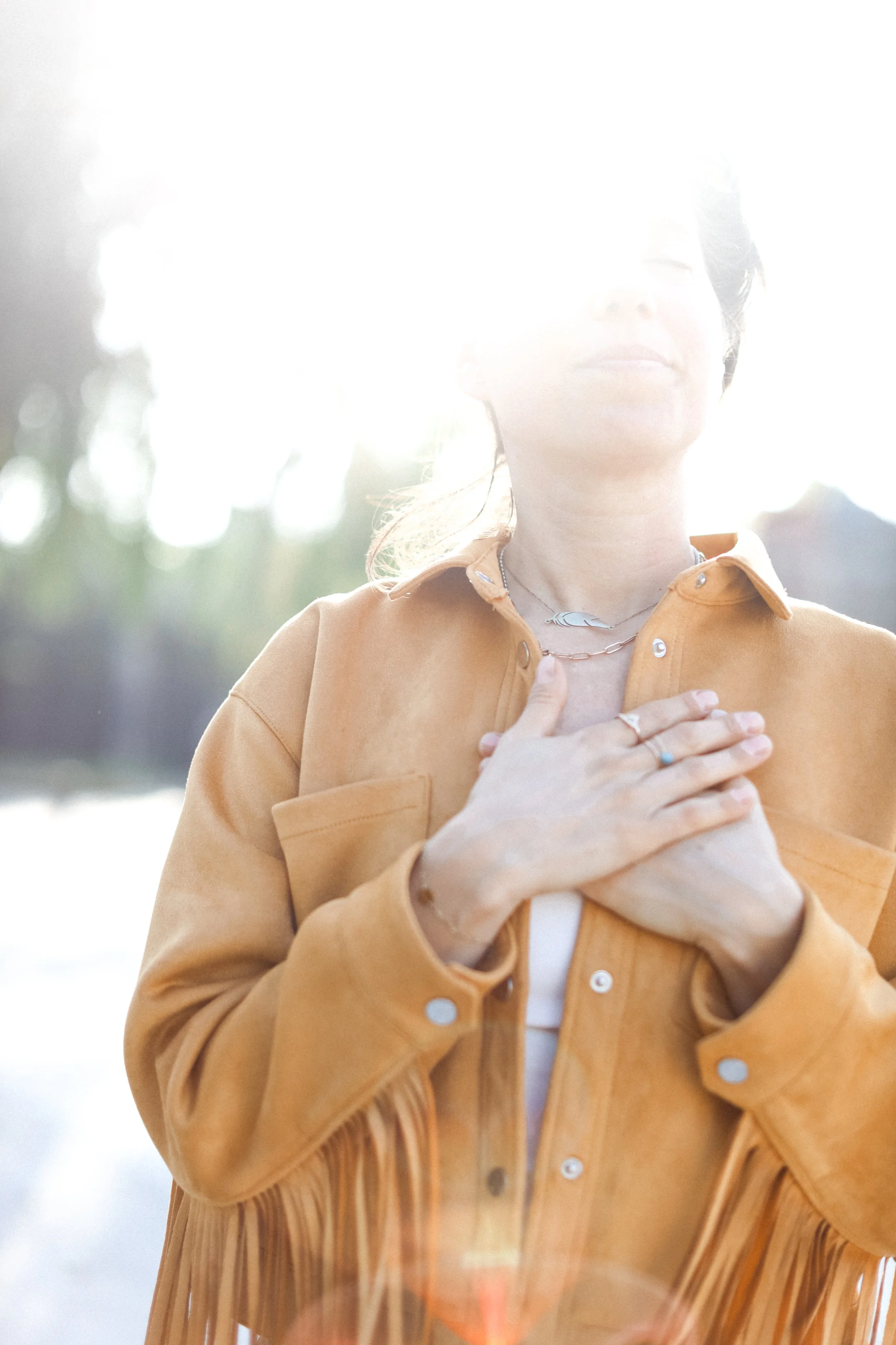 A person standing outdoors with their hands over their chest, wearing a tan fringe jacket and jewelry, backlit by bright sunlight.
