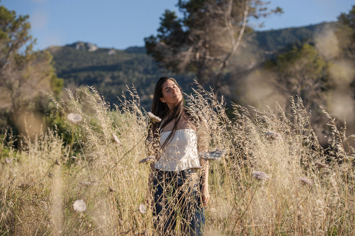 Young woman with long dark hair standing in tall grass and wildflowers in a field, with mountain range and trees in the background on a sunny day.