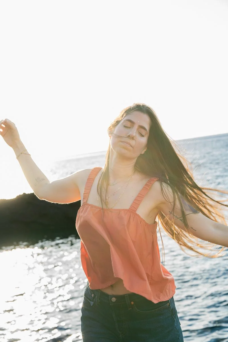 A woman with long hair wearing a flowing orange sleeveless top and dark jeans enjoying herself outdoors near the water during sunset.