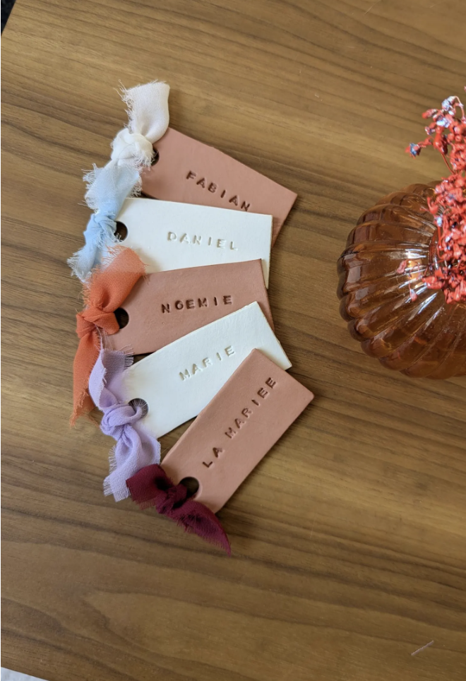 Etiquetas de tela en colores pastel con nombres escritos en ellas, colocadas en una mesa de madera, junto a un jarrón de cristal con flores rosadas.