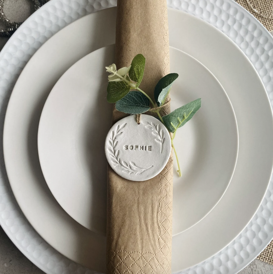 Configuración de mesa formal con plato blanco, servilleta de papel marrón, etiqueta personalizada con el nombre Sophie, decorada con hojas verdes y ramas finas.