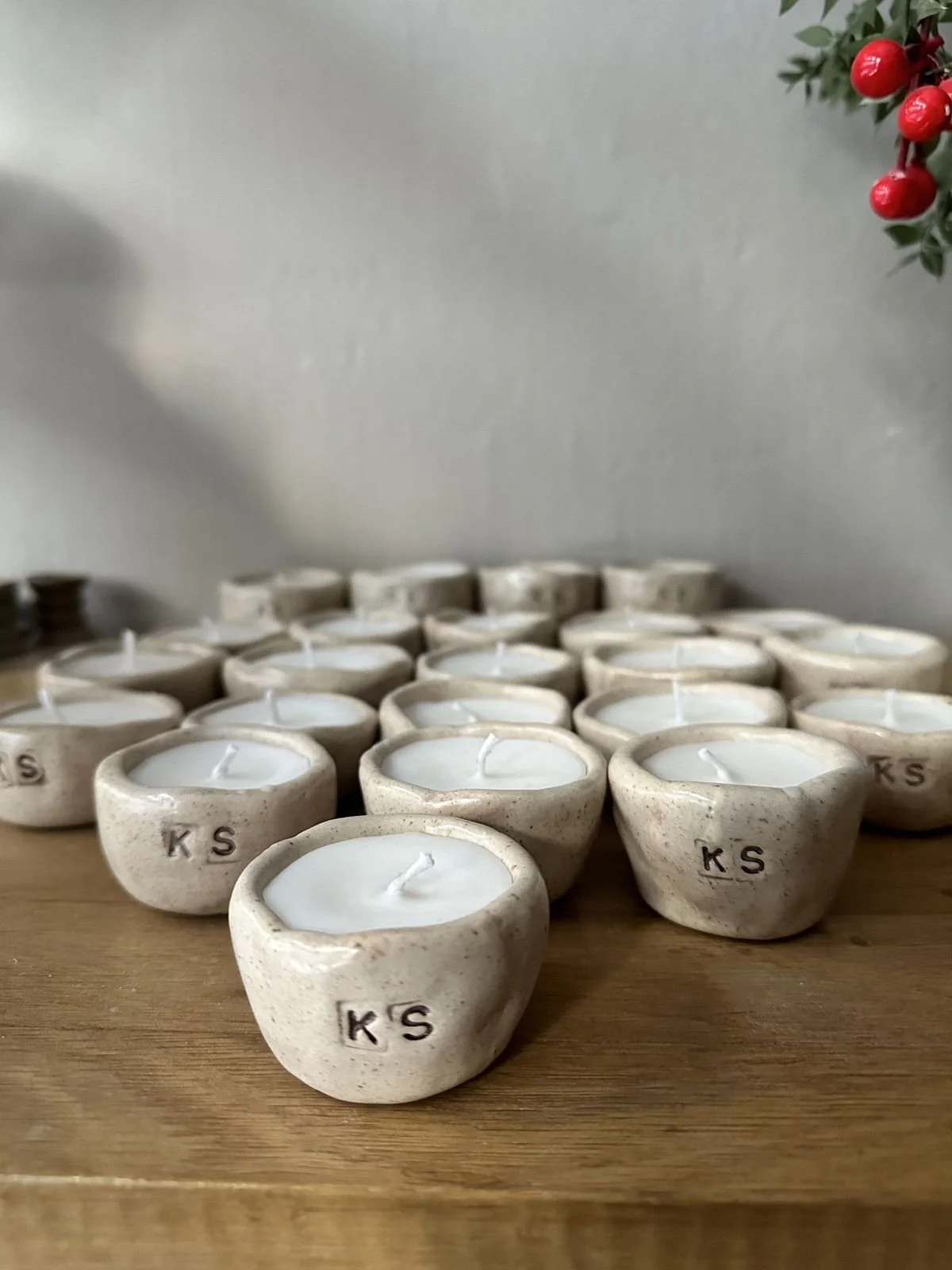 Velas de cera en pequeños recipientes de cerámica con letras 'KS' en una mesa de madera, decoradas con ramas de muérdago con bayas rojas en la esquina.