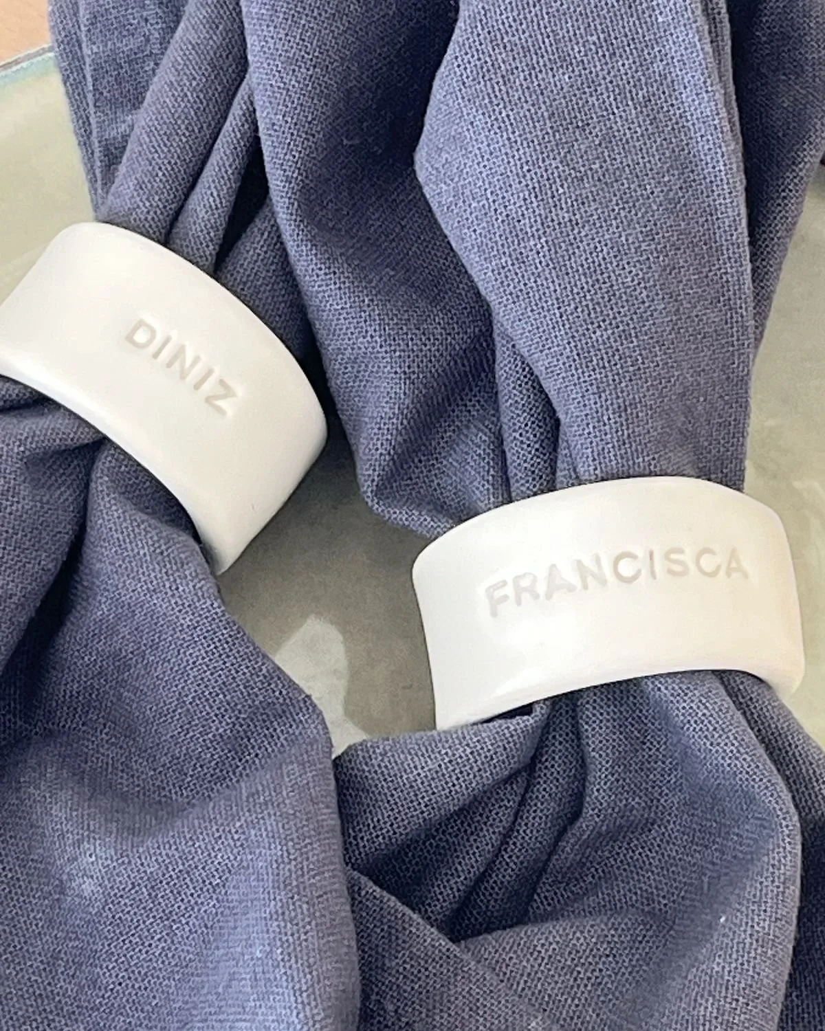 Dos bandas blancas con nombres grabados, uno con 'FRANCISCA' y otro con 'DINZO', colocadas en una tela gris. La tela parece ser de color gris azulado y tiene una textura de punto pequeño.