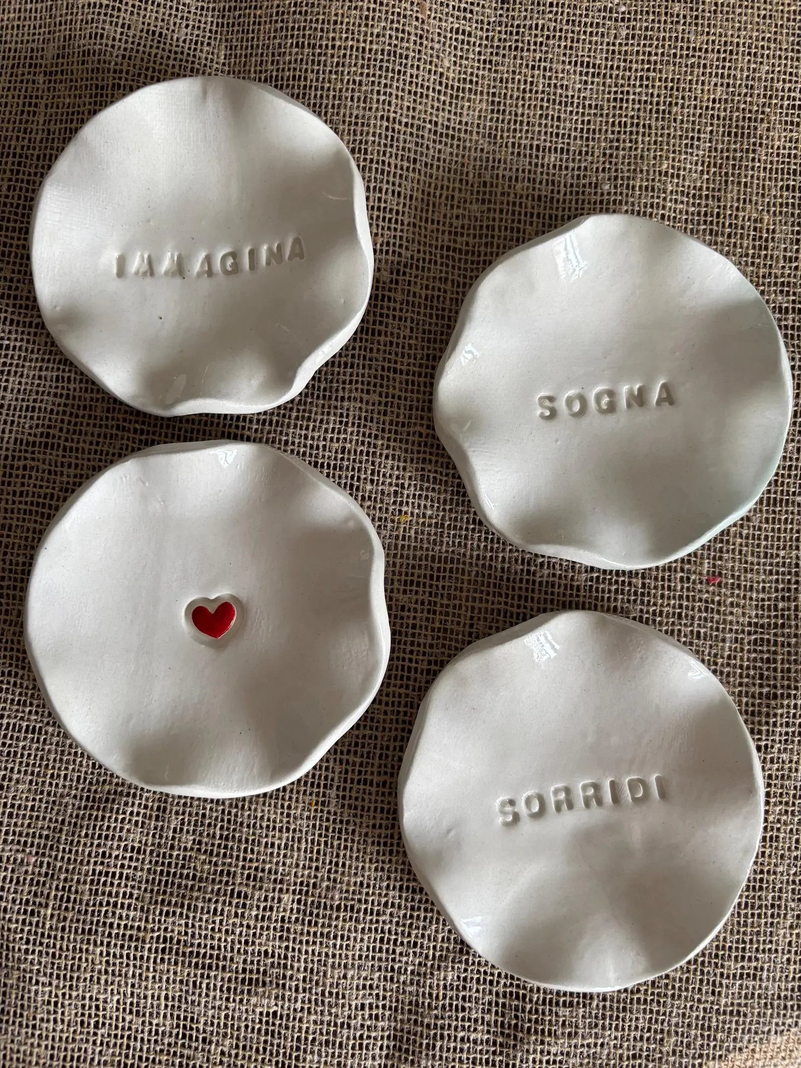 Cuatro platos pequeños de cerámica blanca, dos con palabras en italiano: "SOGNA" y "SIGNORE"; uno con un pequeño corazón rojo en el centro y otro con palabras en italiano: "SORRIDI". Todos están colocados sobre una superficie de tela de color marrón.