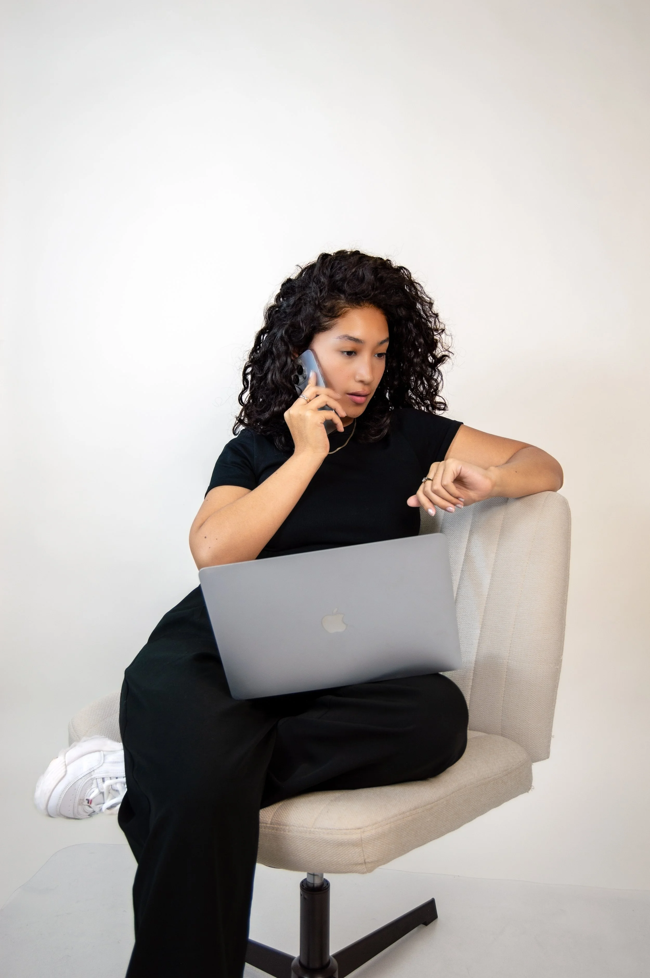 Young woman with curly hair sitting on a beige office chair, wearing a black t-shirt and black pants, talking on a landline phone, with a silver laptop on her lap, against a plain white background.