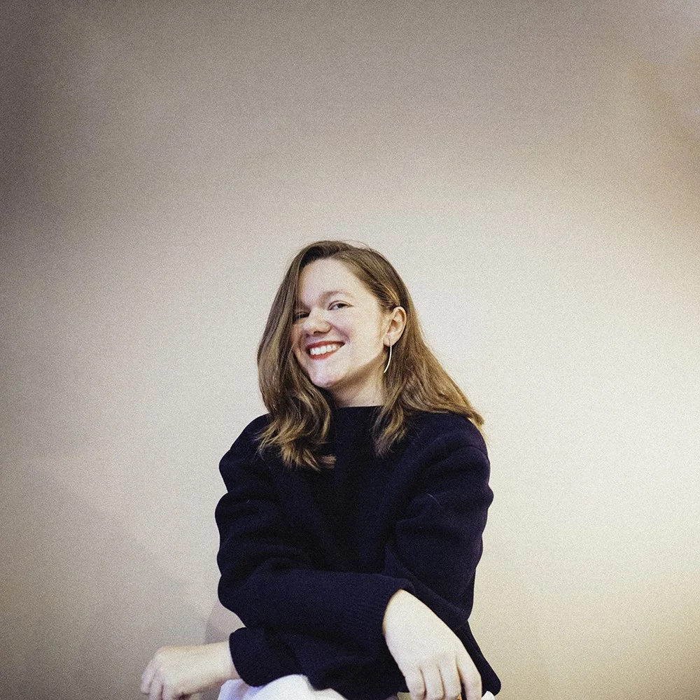 A smiling woman with shoulder-length brown hair, wearing a black sweater, sitting against a plain beige wall.