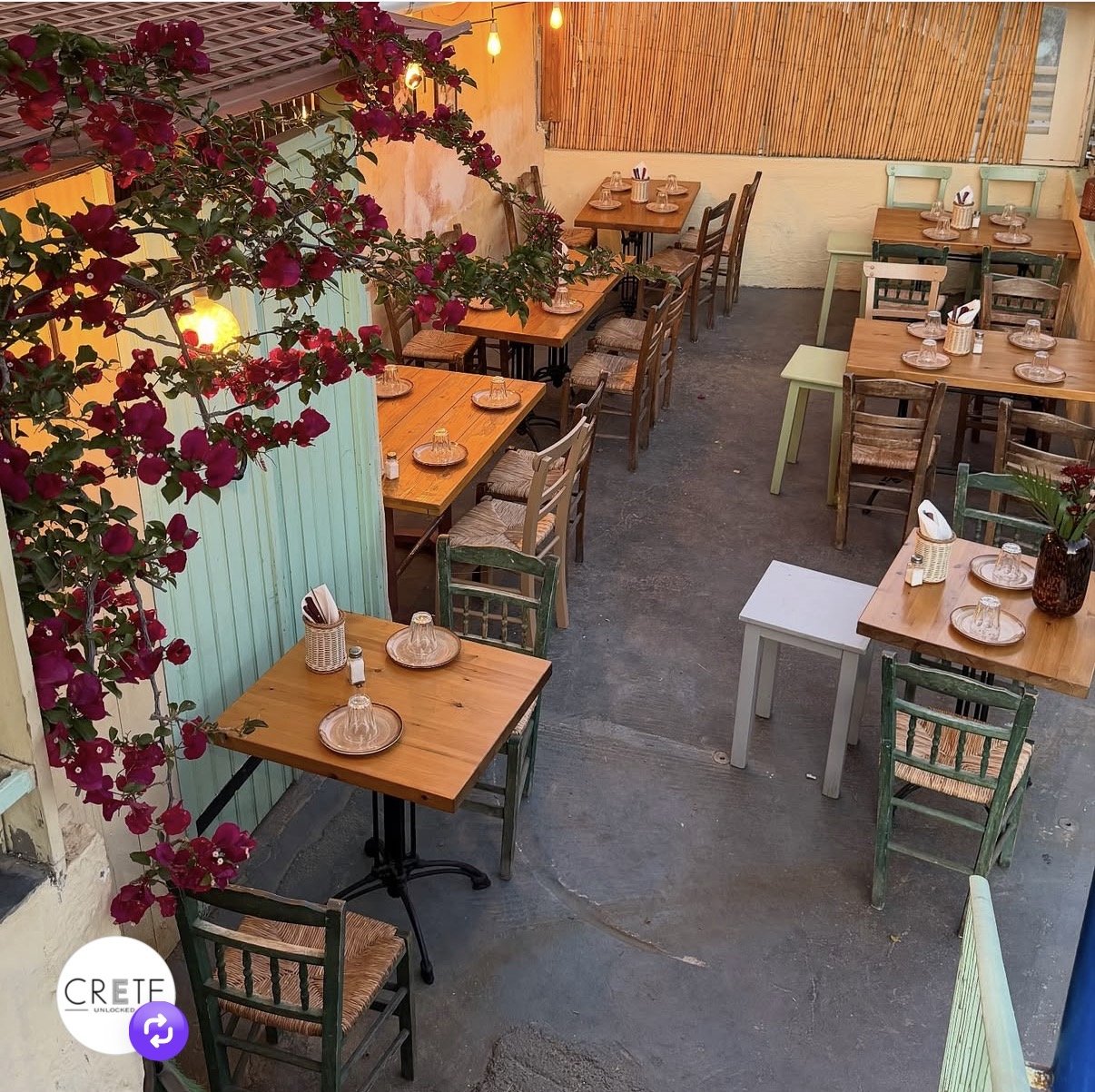 Outdoor Greek restaurant patio with wooden tables, woven chairs, bougainvillea flowers, and warm lighting