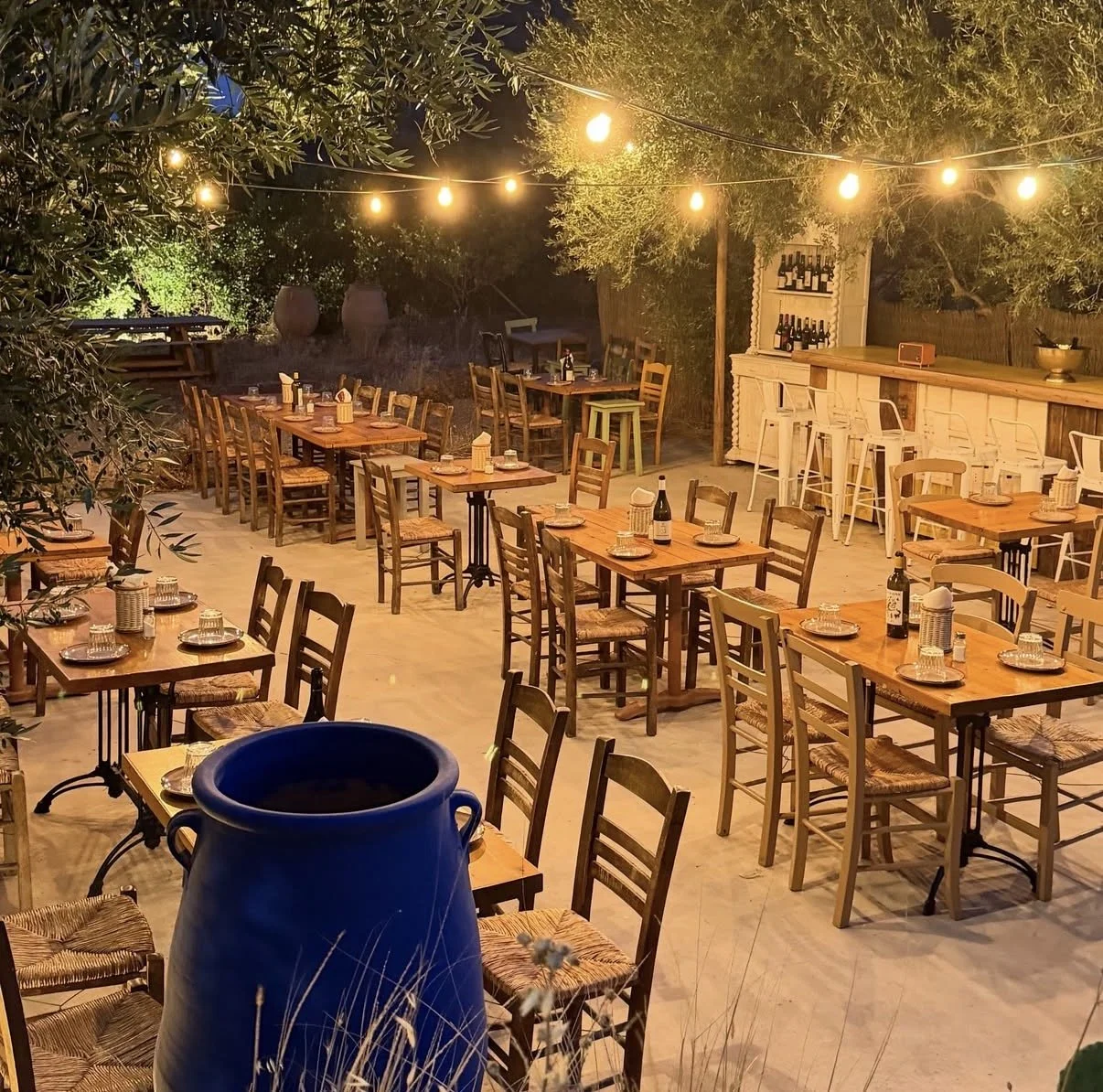 Outdoor restaurant patio at night with wooden tables, chairs, string lights, and a bar surrounded by trees.