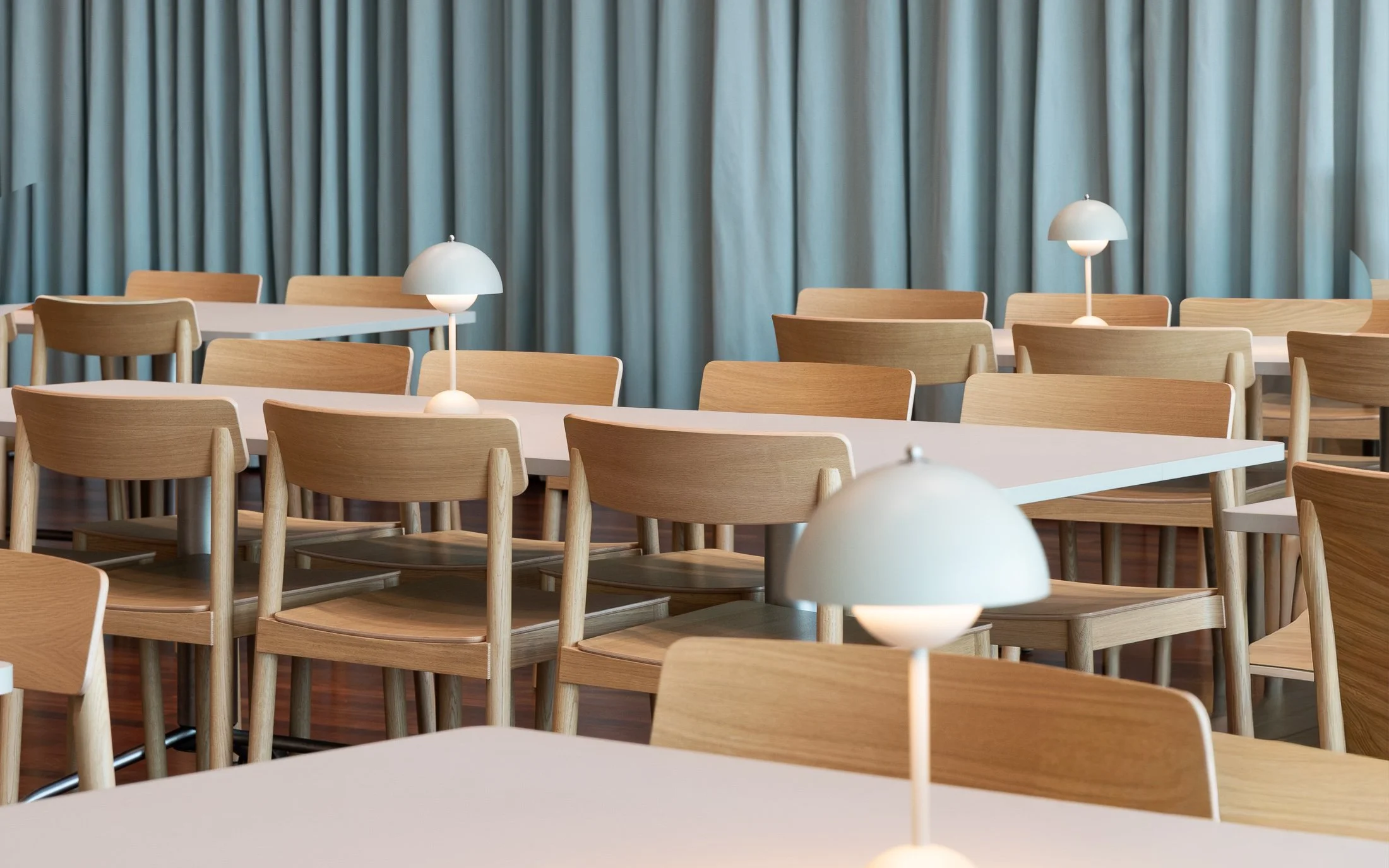 A minimalist dining area with a beige bench, two wooden chairs, a small white table lamp, and a wooden dining table, set against a backdrop of light blue curtains.