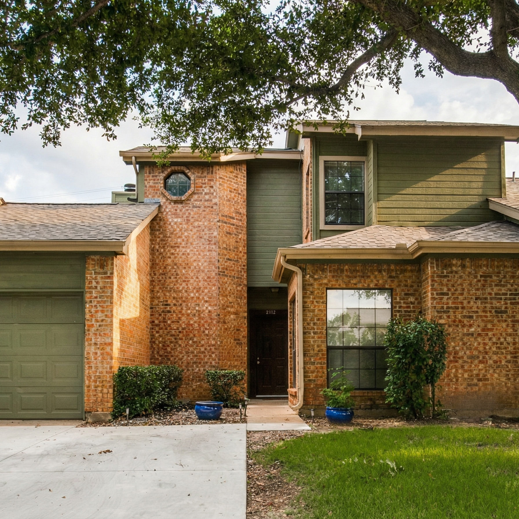 Front view of a two-story house with brick and green siding, a large window, and a brown door, with a small front lawn and potted plants in blue pots.