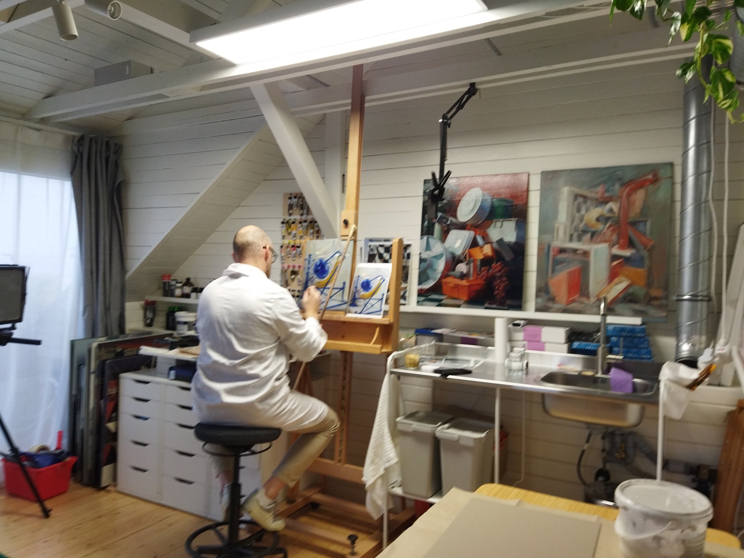 An artist in white clothing painting abstracts at an easel in a well-lit art studio with paintings on the wall and art supplies around.