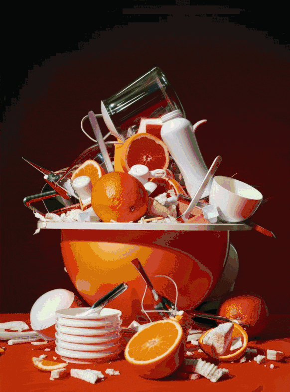 Broken dishes, glasses, and shattered orange slices spilled on a table with a large orange bowl filled with broken porcelain and zest, against a dark background.