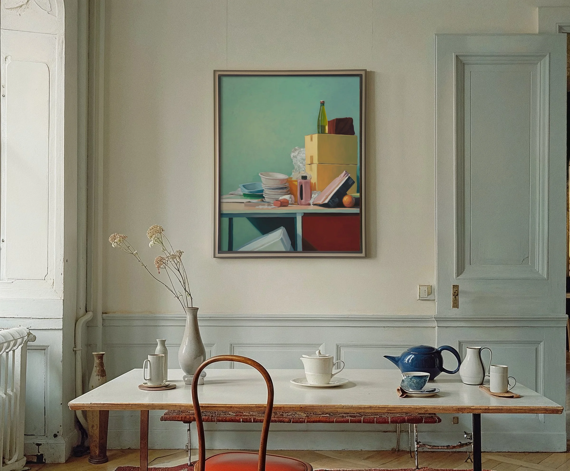 A dining room with a wooden table set with various dishes, a blue teapot, and a chair. On the wall hangs a framed still-life painting of kitchen items, and a vase with dried flowers sits on the table.