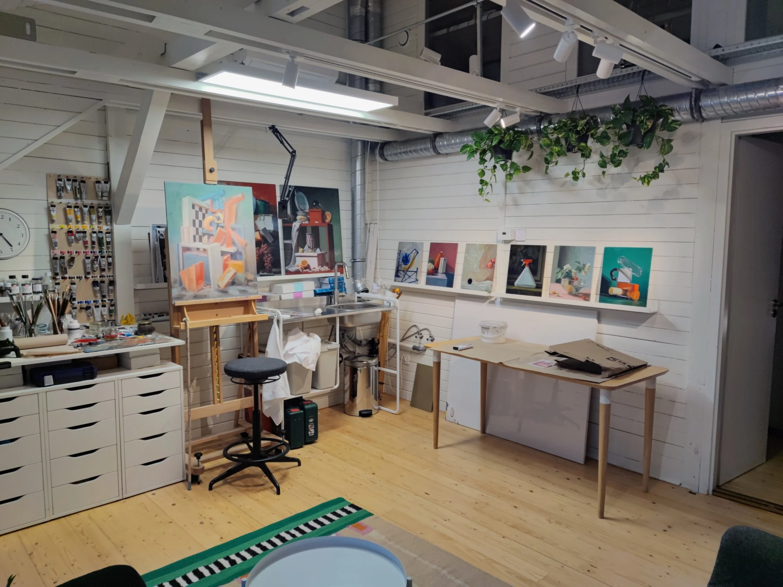 An art studio with paintings on the wall, an easel, a stool, and various art supplies, with white wooden walls and ceiling, and green hanging plants.