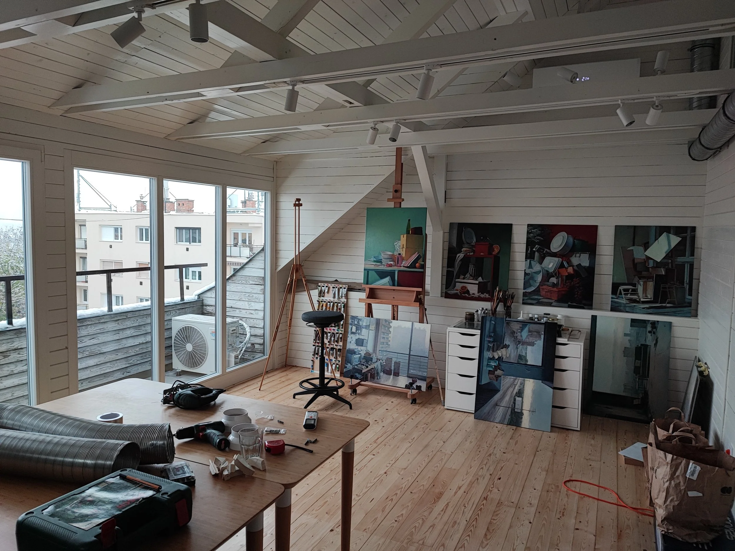An artist's studio with large windows, artist's easels and paintings, a wooden table with painting supplies, and various art works in progress.
