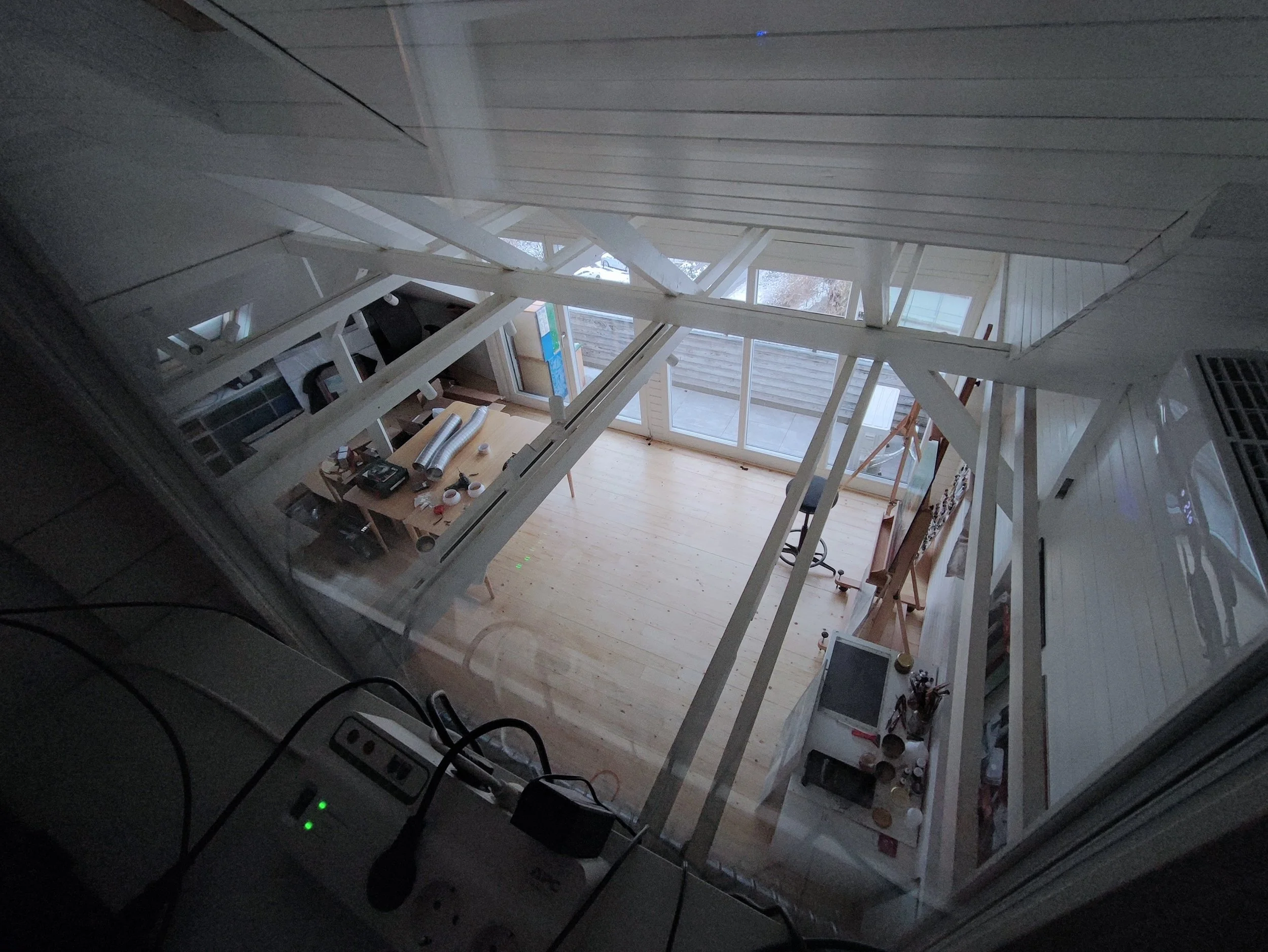 Looking down through a staircase into a sunlit living area with large sliding glass doors, a wooden floor, and a table with various items on it, including a phone, rolls of material, and small objects, viewed from an upstairs window.