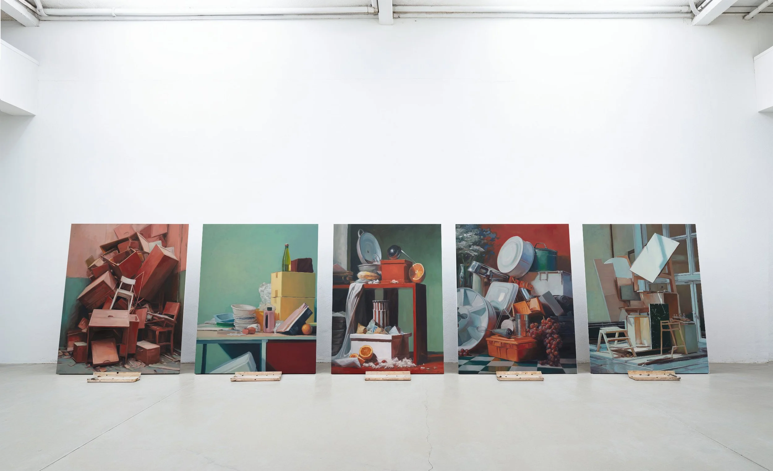 Five large paintings displayed on wooden pallets in a white gallery space, depicting various still life scenes with objects like chairs, household items, and food.