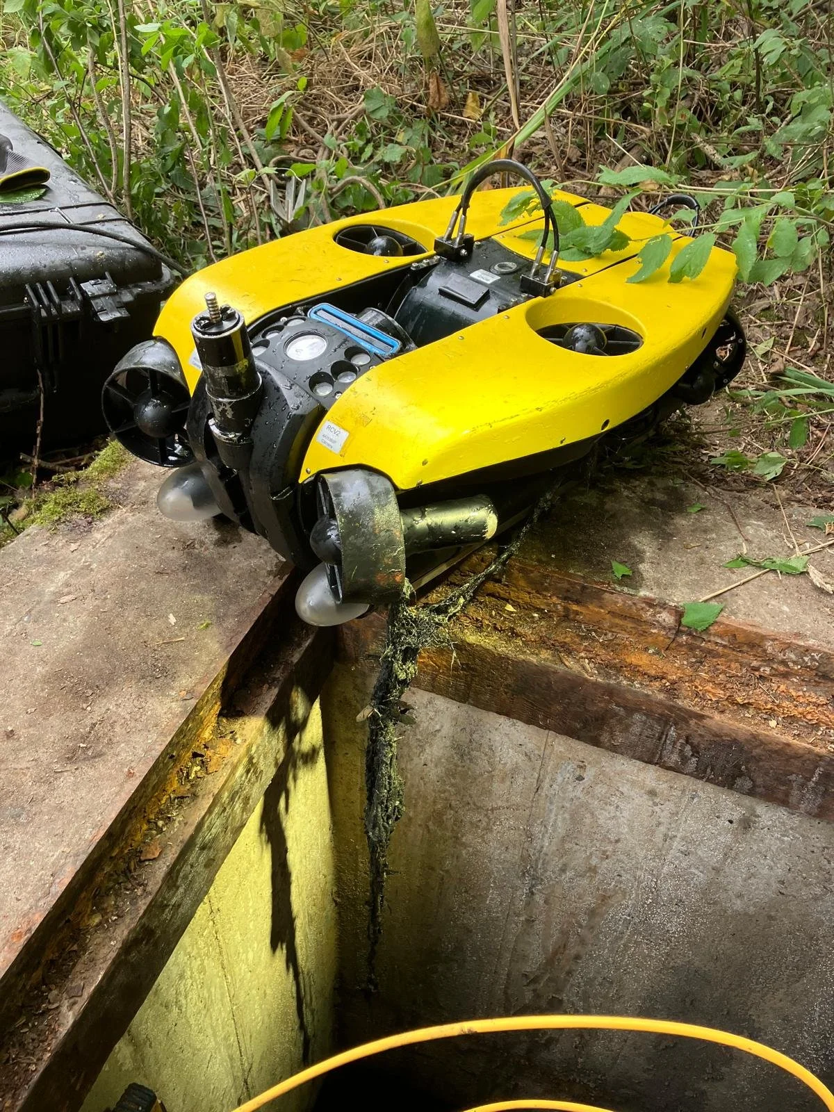 A yellow underwater robot or drone with propellers on each side, lying on a concrete and wood surface in a natural, outdoor setting with greenery and bushes.