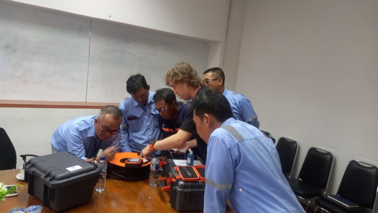 Group of men wearing blue work uniforms gathered around a table examining technical equipment in a conference room.