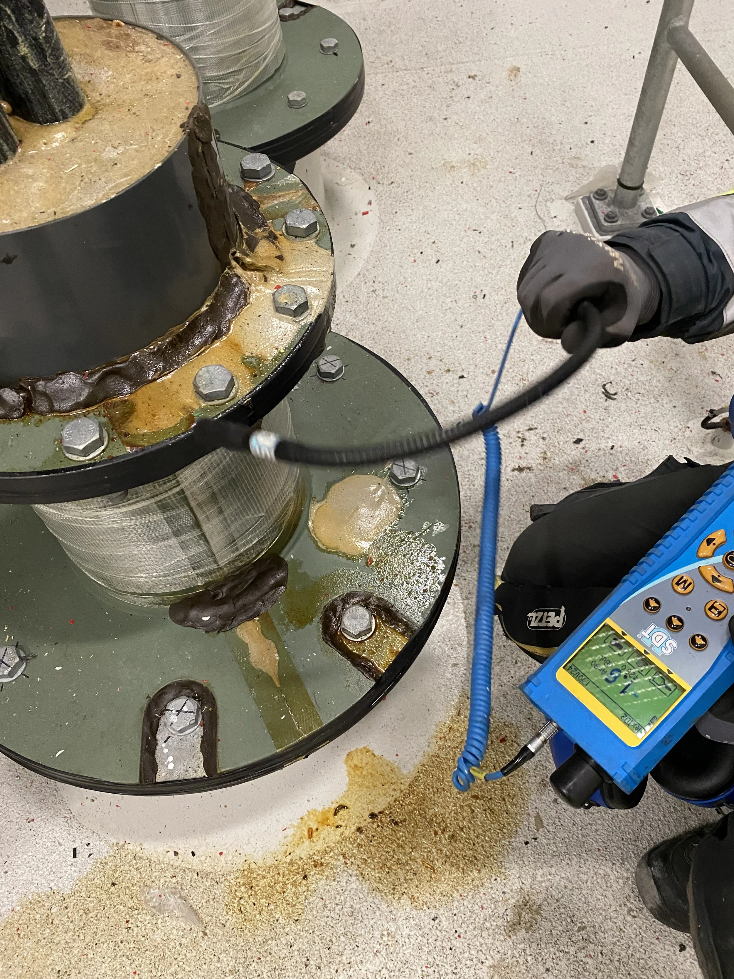 A person inspecting a large industrial equipment part with a handheld instrument, possibly a ultrasonic or thickness gauge, in a workshop or factory setting. There are visible grease, oil, and debris on the floor.