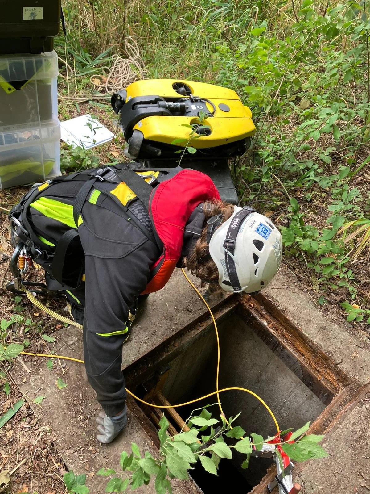 A person wearing a helmet and rescue gear kneels beside an open underground utility or sewer access point. They are inspecting or working inside the rectangular opening, which is surrounded by dirt and vegetation. Various tools and equipment are nearby, including a yellow rescue board and a plastic storage container.