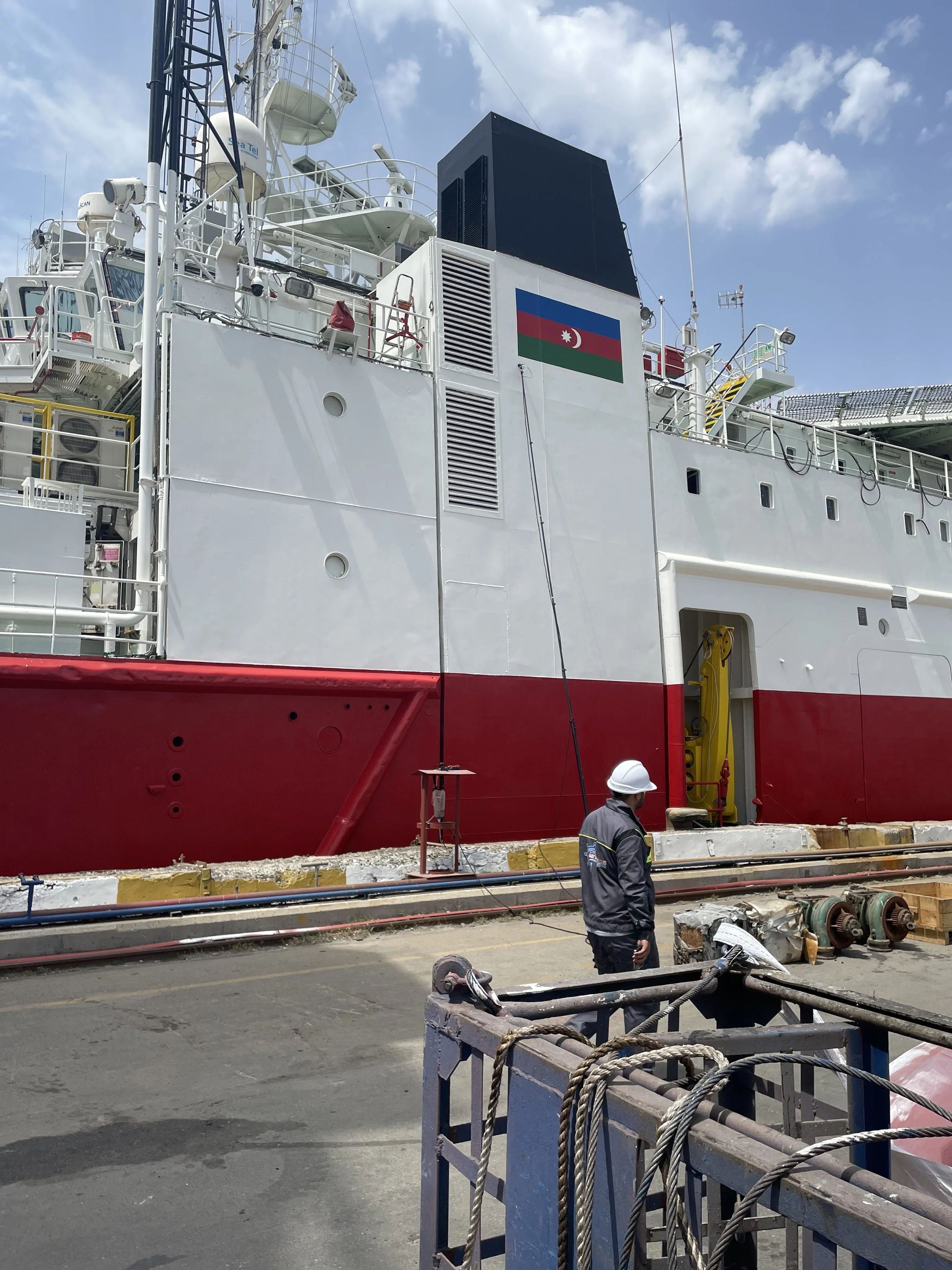 A large vessel painted white with red at the bottom, with a banner of the Azerbaijani flag on it, docked at a port. A person wearing a white helmet and dark work clothes stands nearby, and there are industrial equipment and pipes on the ground.