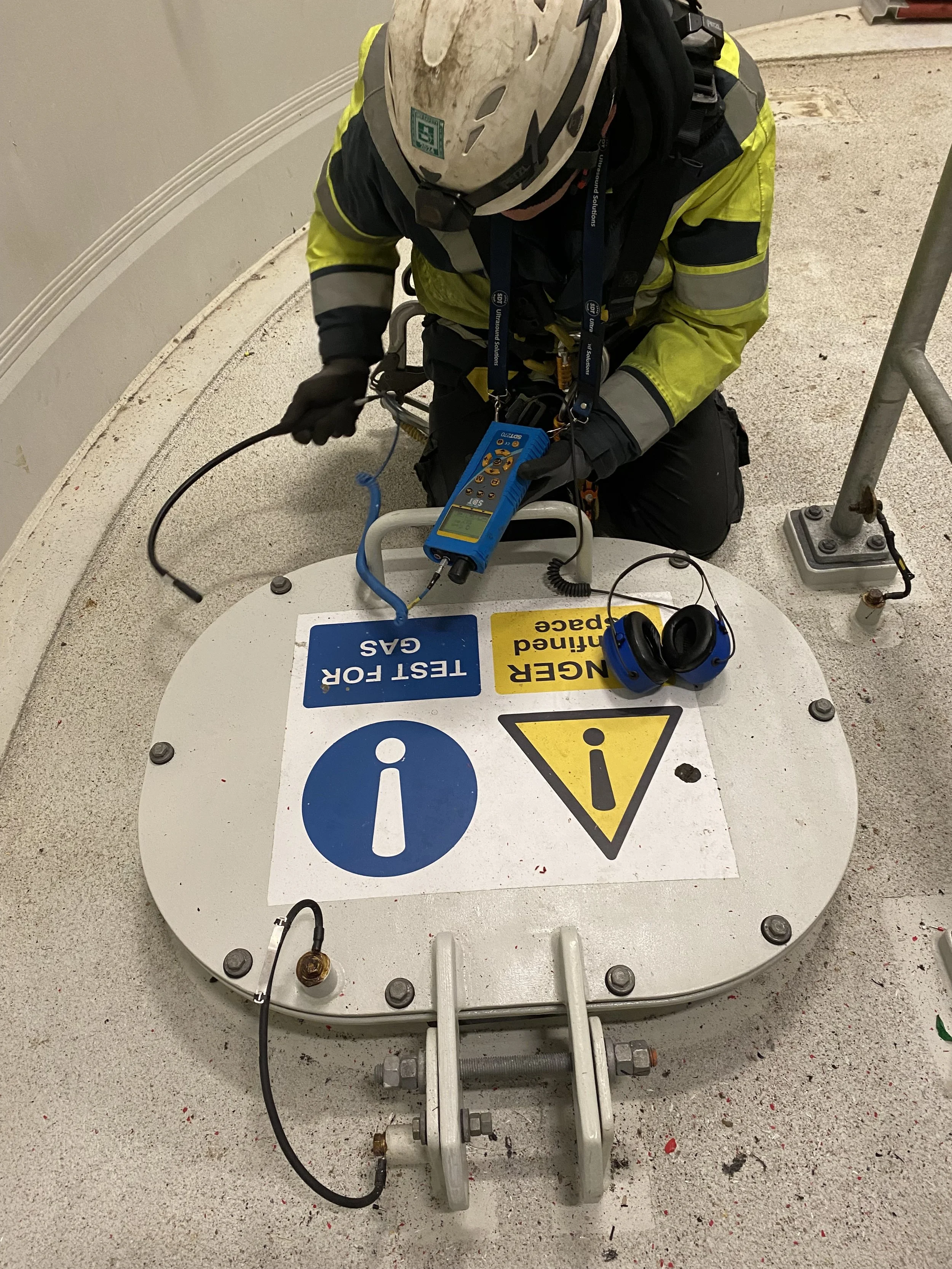 A worker in safety gear, including a helmet and gloves, kneeling on a concrete surface, working with electrical and safety equipment on a raised platform. The platform has warning and information signs, including caution symbols and instructions.
