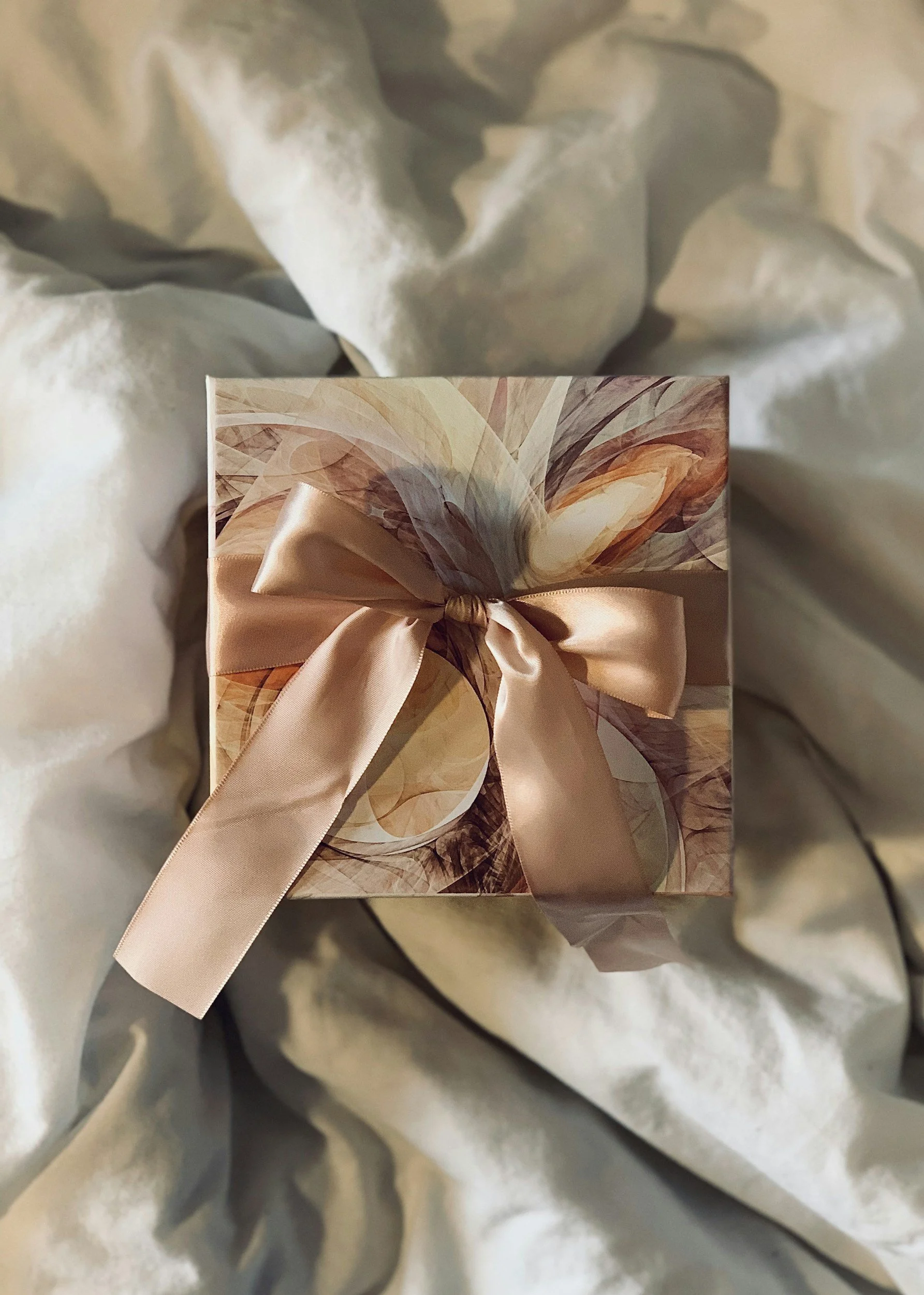 A gift box wrapped in abstract-patterned paper with a satin ribbon tied into a bow, resting on crumpled white tissue paper.
