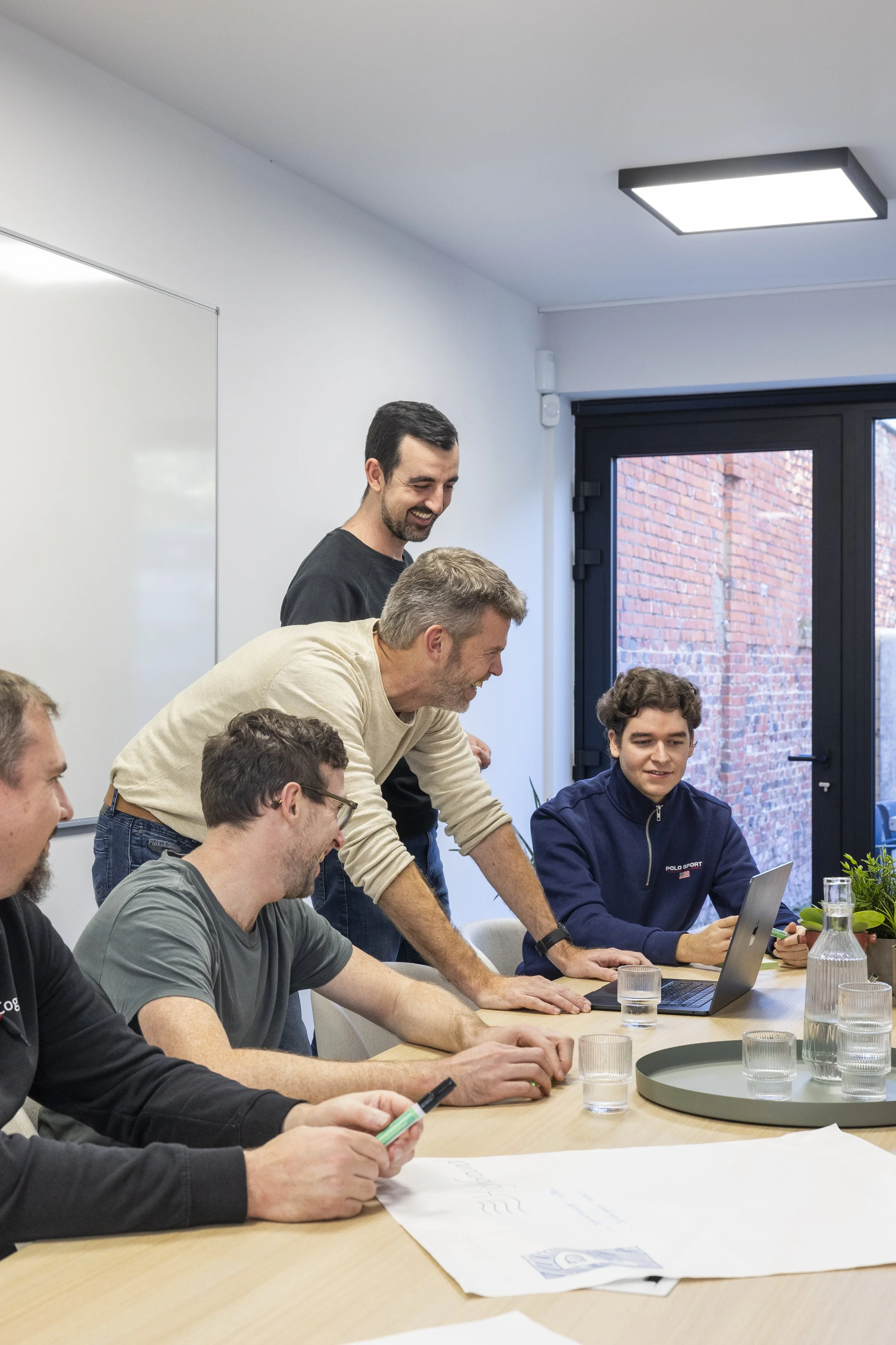 Vijf mensen die samen lachen en werken rond een tafel in een kantoor, met een laptop en papieren.
