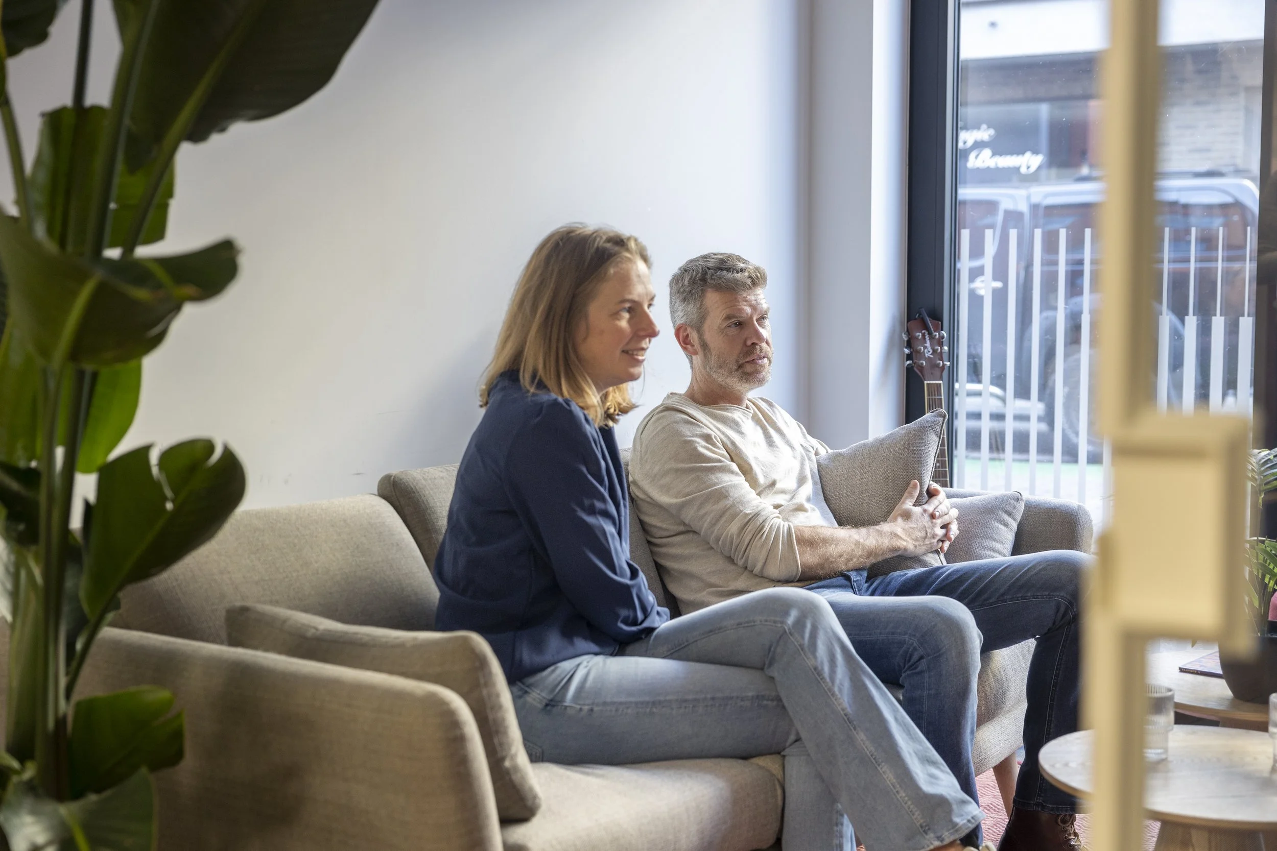 Twee mensen zittend op een bank in een woonkamer, met een groot raam en een gitaar in de achtergrond.