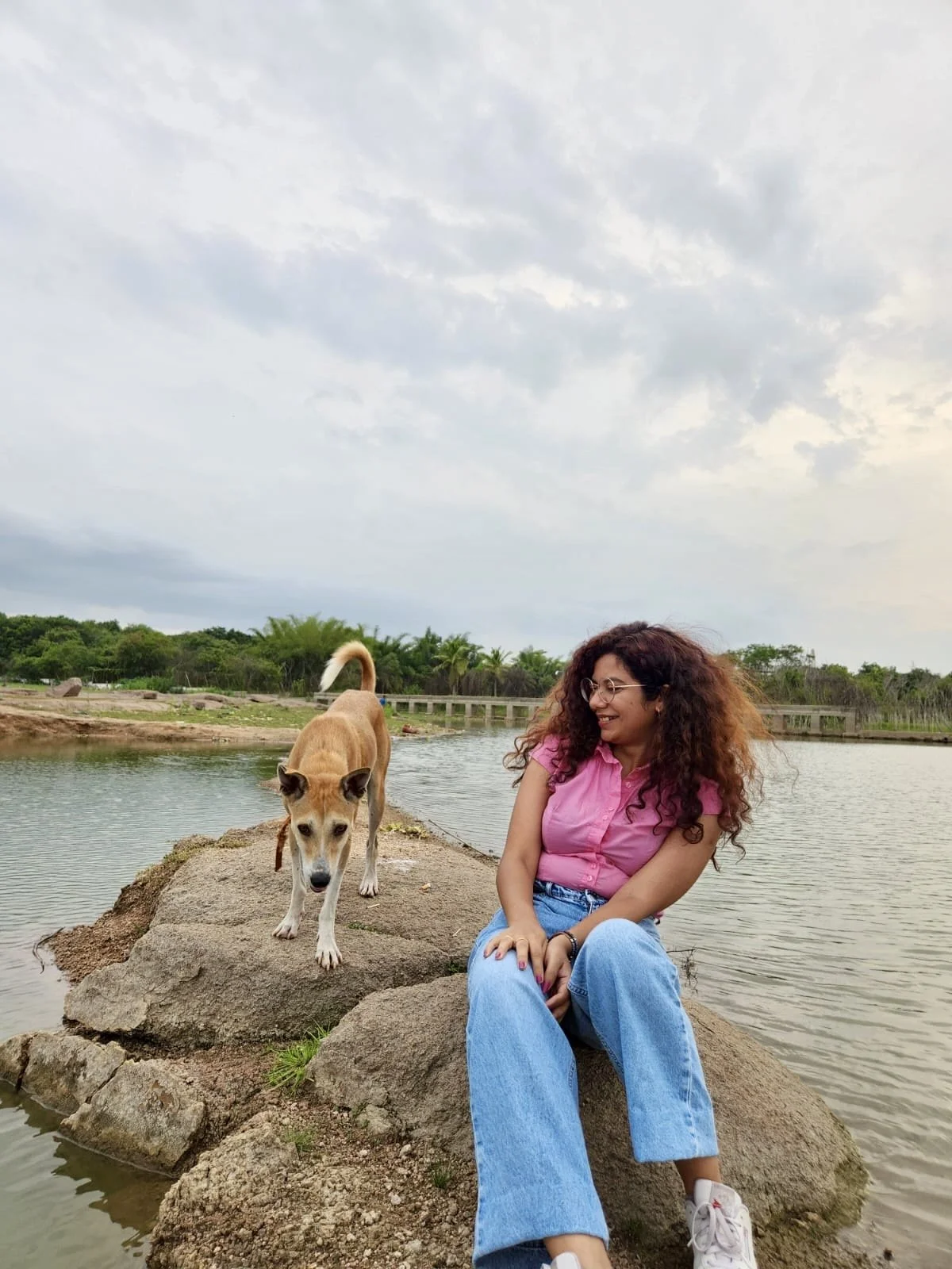 A young woman with curly hair wearing glasses, a pink shirt, and jeans sitting on a large rock near a body of water, smiling while looking at a tan dog standing nearby on the same rock.