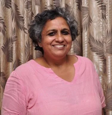 A woman with short curly gray hair, wearing a pink shirt, smiling in front of a patterned curtain.