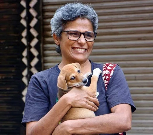 An older woman with gray hair, glasses, and a navy T-shirt smiling while holding a small tan and white puppy outdoors.