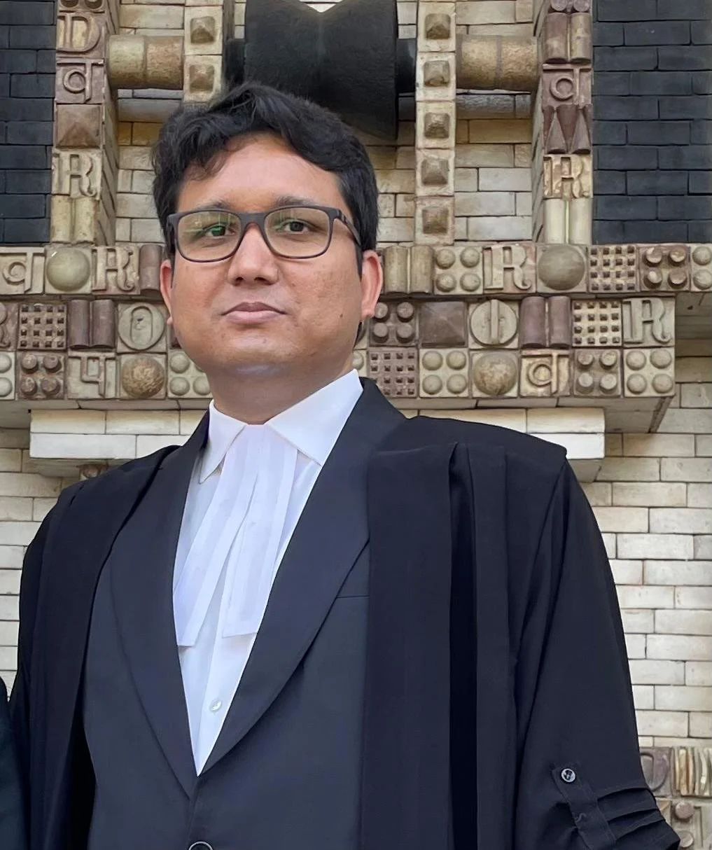Close-up of a man wearing academic graduation robes and glasses, standing in front of a decorative brick wall with symbols and patterns.