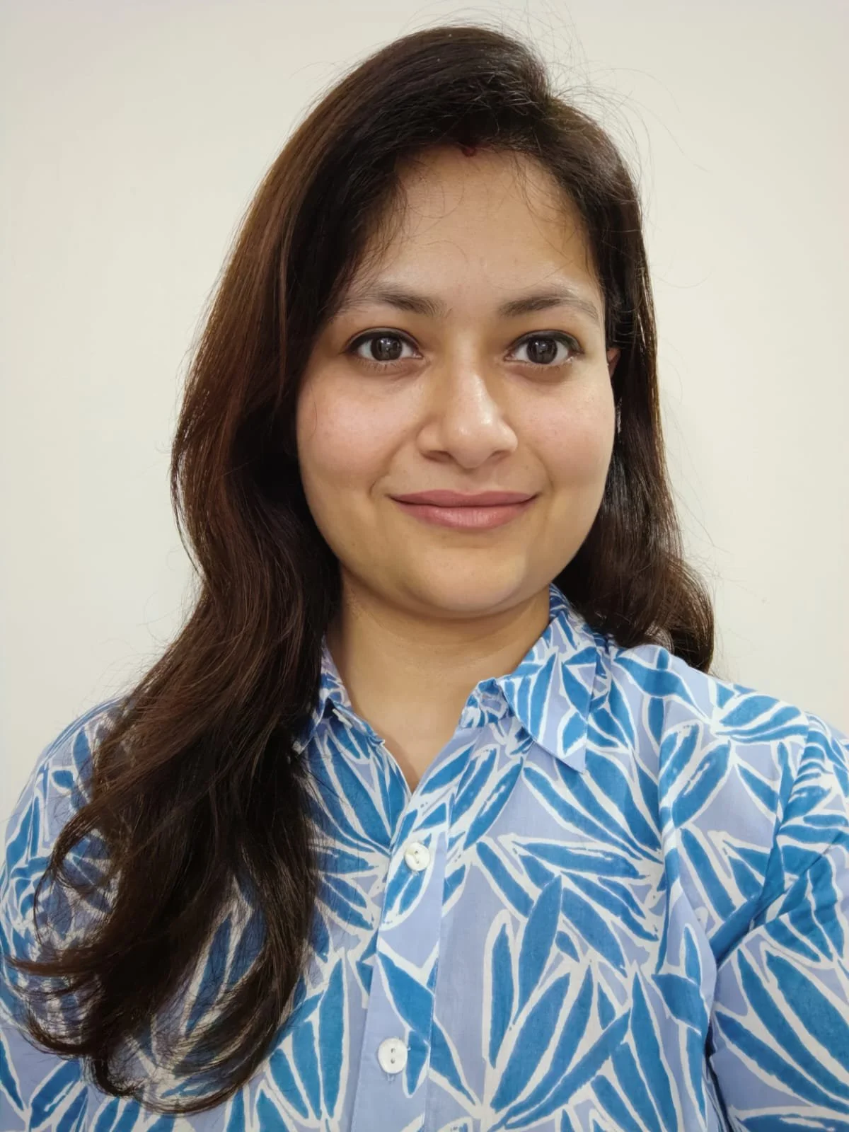 A woman with brown hair wearing a blue and white patterned shirt, standing against a plain light background.