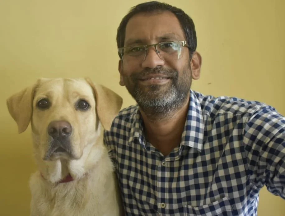 A man with glasses and a beard smiling for a selfie with a yellow Labrador Retriever against a plain yellow wall.