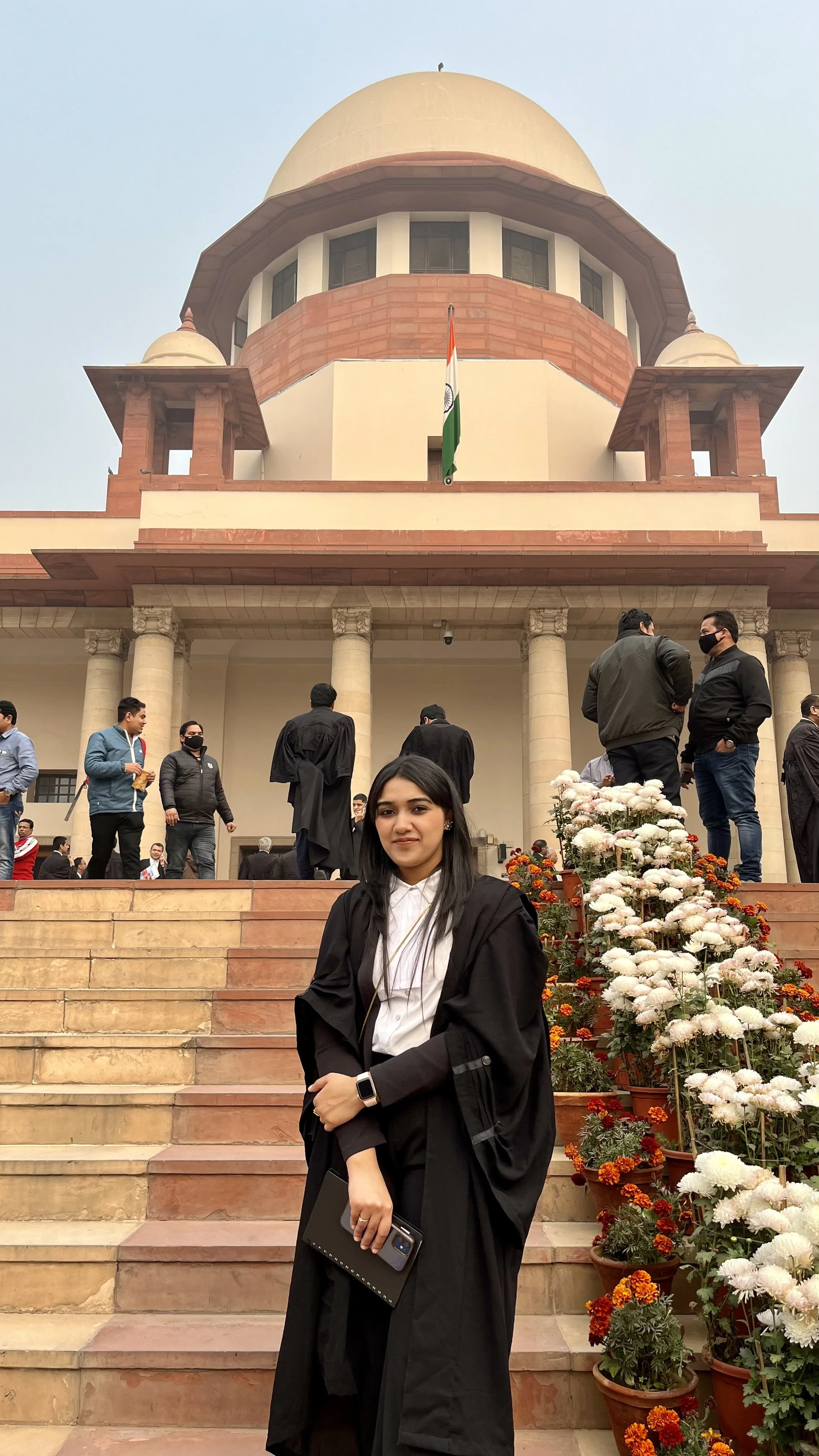 A young woman in a graduation gown standing on the steps of a government building, with other people in the background, some wearing masks, and flowers along the stairs.