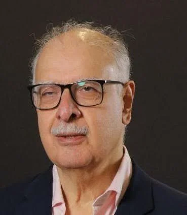 A portrait of an elderly man with glasses, wearing a dark blazer and light pink shirt, against a dark background.