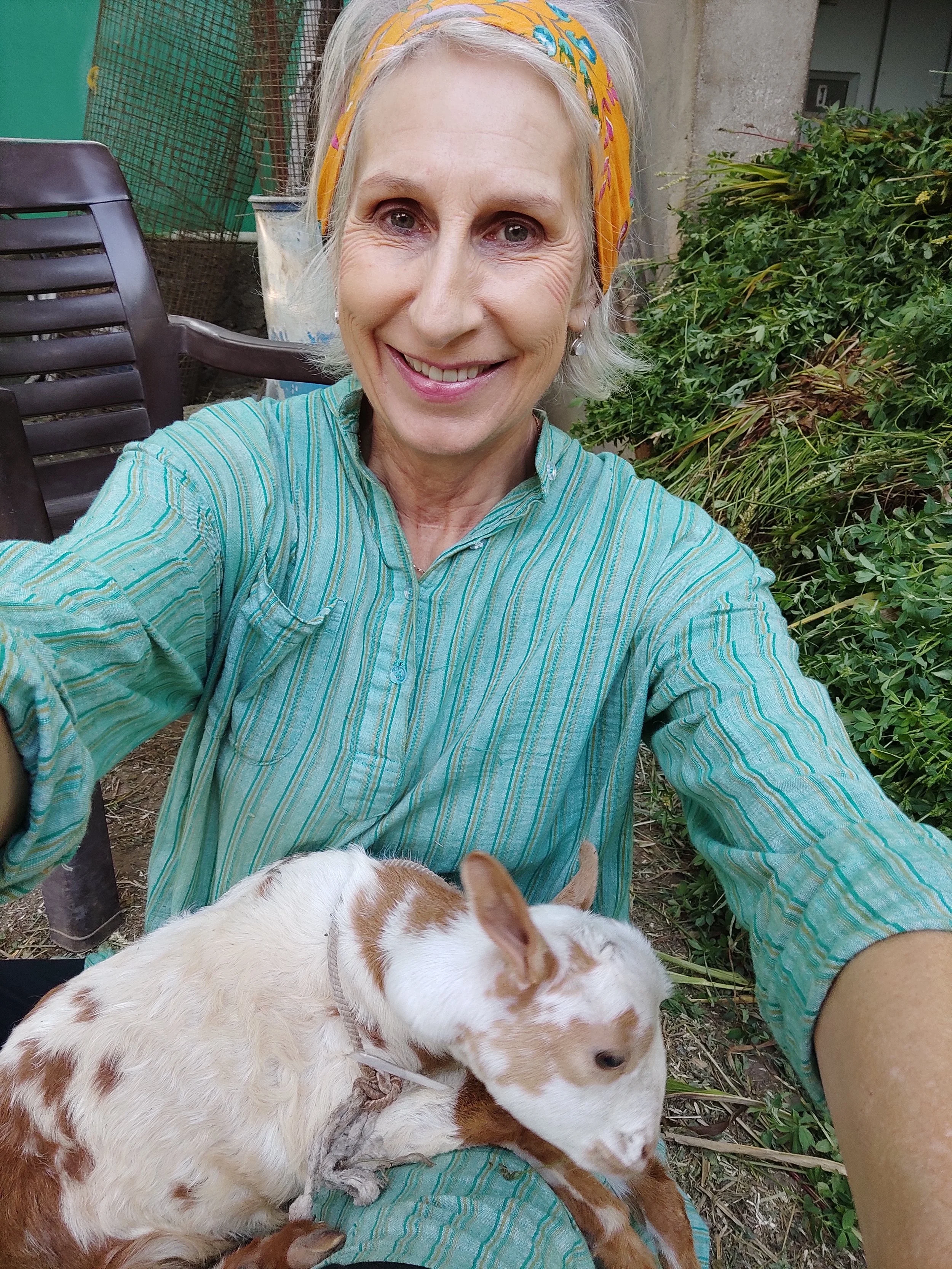 A smiling woman with gray hair wearing a colorful headscarf and a striped turquoise shirt taking a selfie outdoors. She is holding a small goat with white and brown fur, which is resting its head on her lap. She is sitting on a chair next to green bushes and other outdoor furniture.
