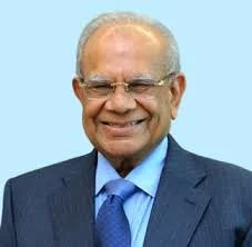 A smiling elderly man with glasses wearing a dark suit, blue shirt, and tie against a light blue background.