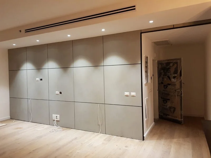 חדר פנים מודרני עם קיר של פאנלים אפורים ותאורה שקועה, הכולל שקעי חשמל וכבלים תלויים. בחדר ריצוף עץ בהיר ופתח פתוח חלקית המוביל לחלל נוסף, עם דלת מכוסה בחומר מגן.