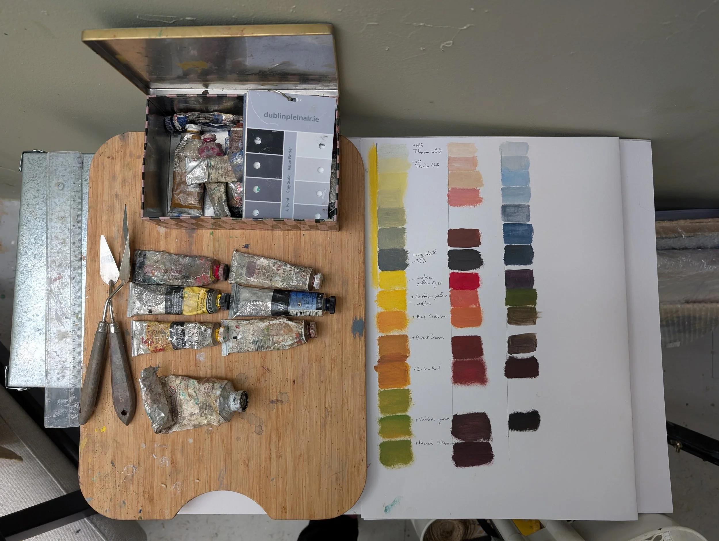 A wooden table with paint tubes, two palette knives, a metal box of paints, and a notebook showing a color palette with various shades, names, and notes.