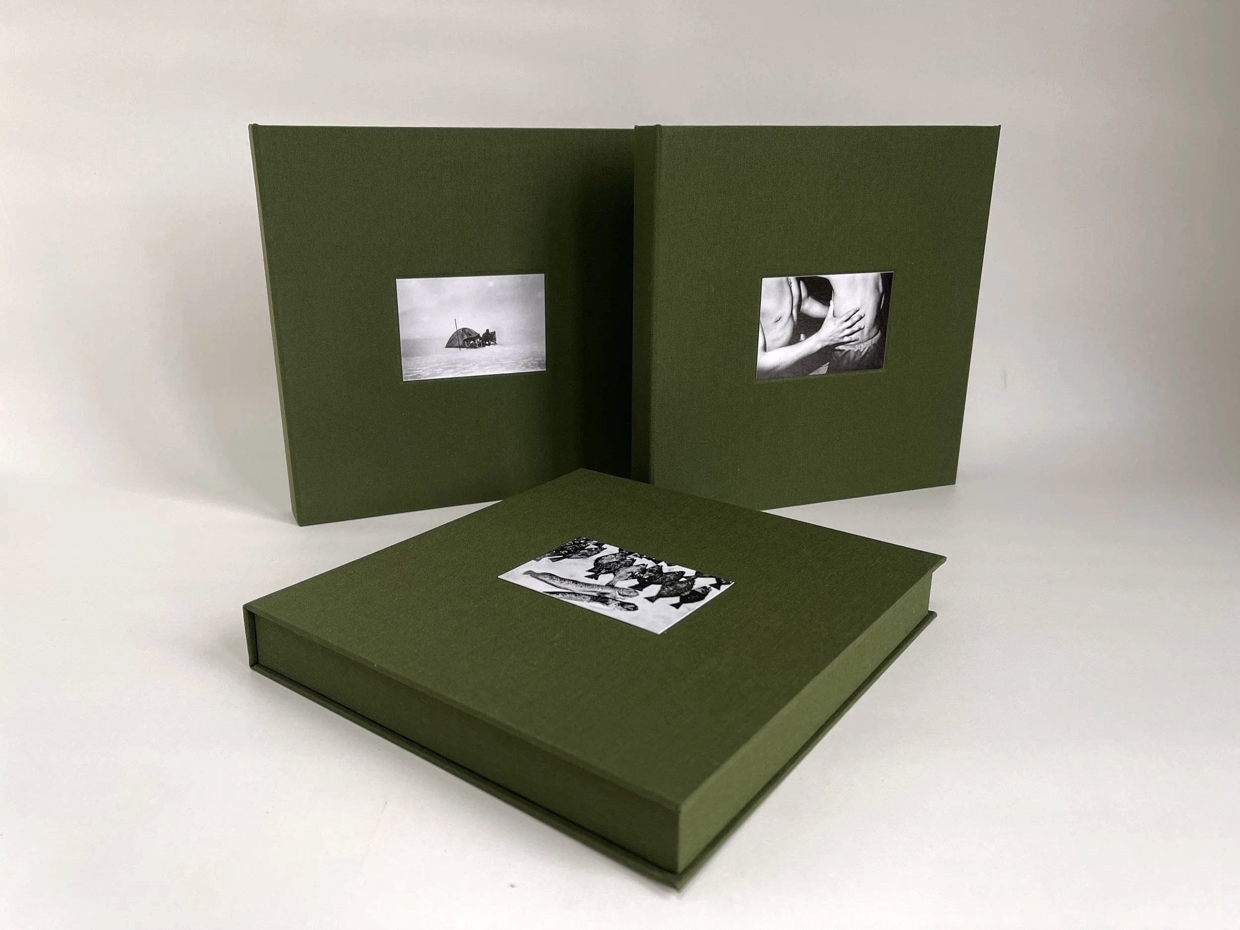A green photo album with three black-and-white photographs inside. One open album showing two photos, one of a boat and the other of a person touching another person's back, and a closed album with a single photo on top of it.