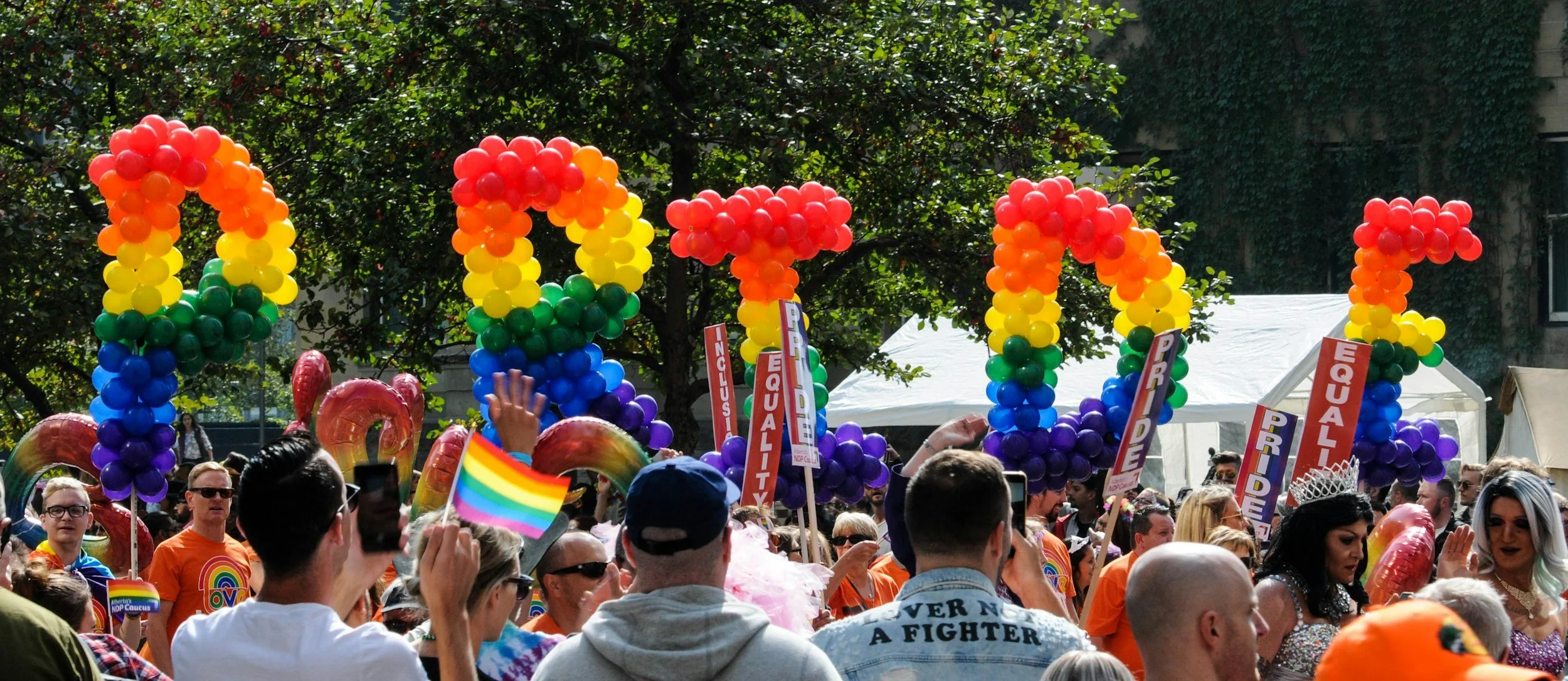 People participating in a pride parade, holding rainbow flags and signs that read, "Inclusion," "Equality," "Pride" and "Respect." There are rainbow balloons spelling out a message, and a person wearing a crown and a rainbow-colored outfit. The crowd is diverse, with some individuals wearing sunglasses and colorful clothing, amidst trees and tents.