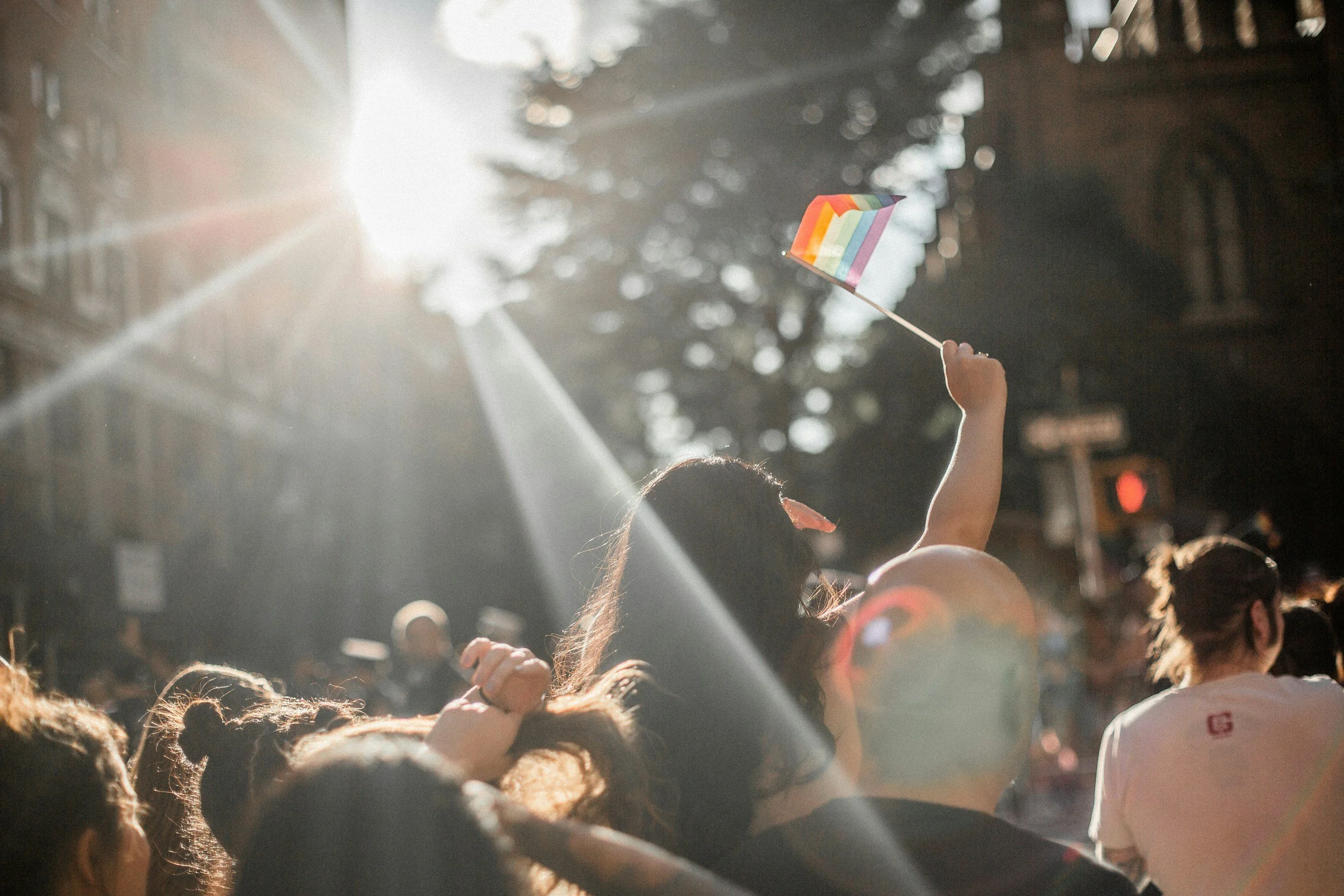 Person holding a rainbow pride flag at an outdoor event during sunset with a crowd in the background.