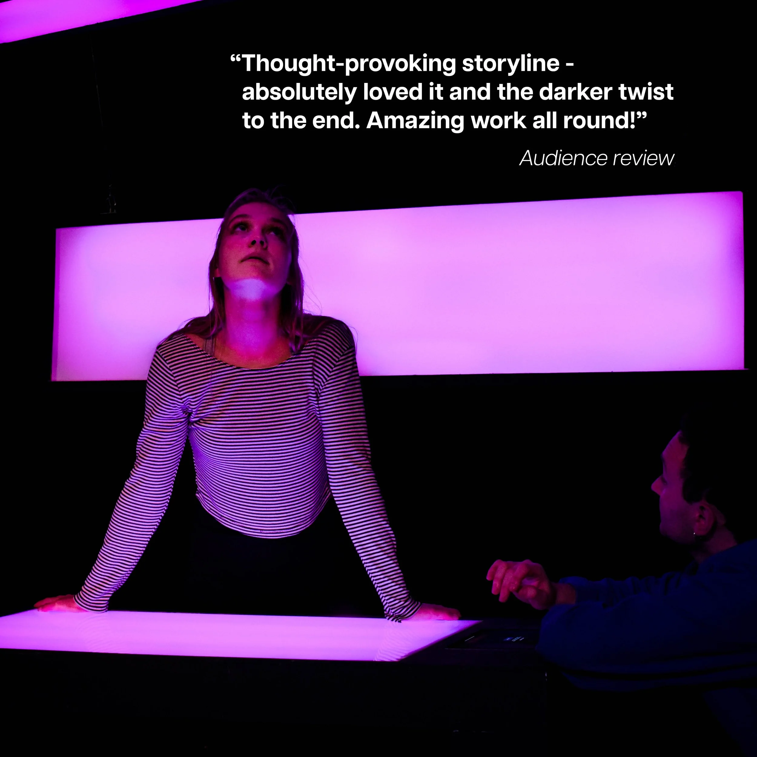 A woman with blonde hair, wearing a striped long sleeve shirt, leaning on a brightly lit table, looking upwards. A man sits across from her, pointing. A quote on a black wall behind them reads: "Thought-provoking storyline – absolutely loved it and t