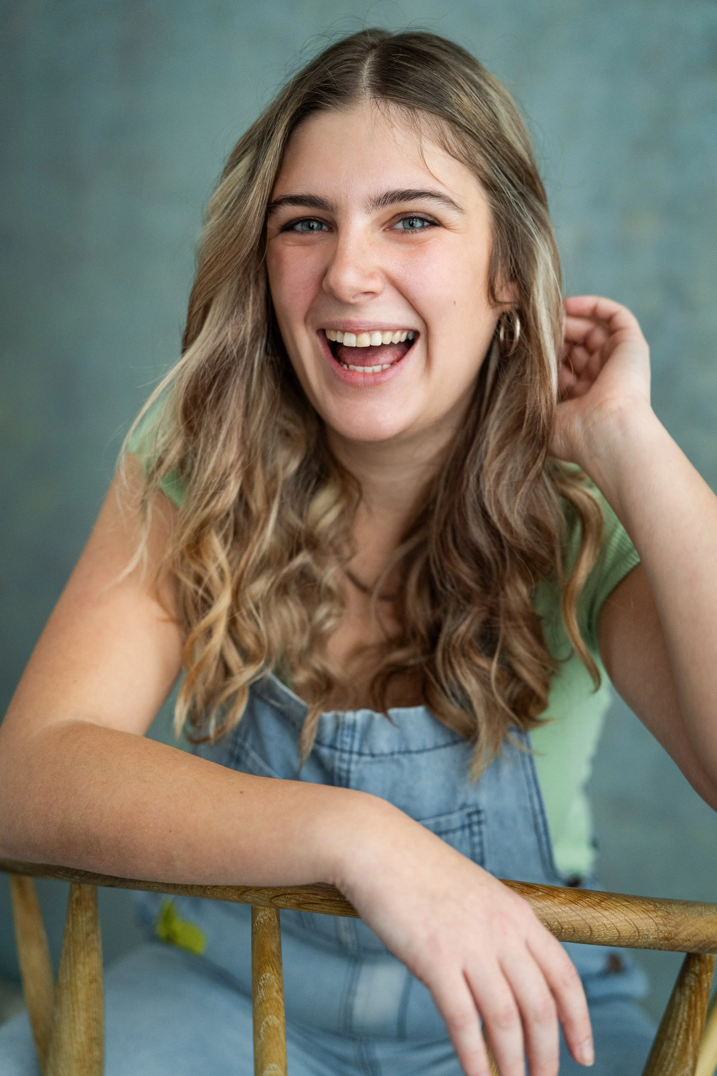 A young woman with long wavy hair smiling at the camera, wearing a green shirt and denim overalls, with one hand touching her hair and leaning on a wooden chair.