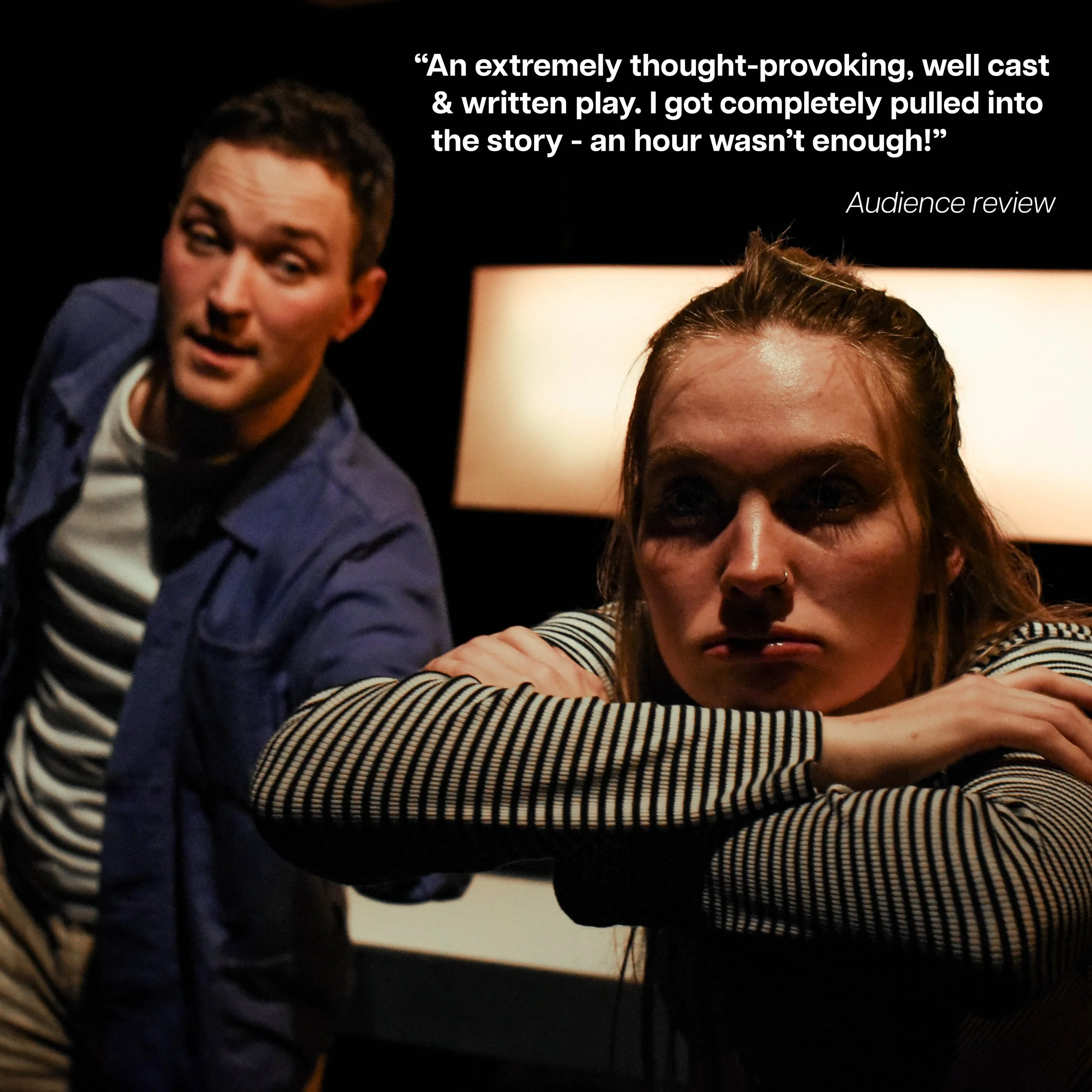 A man and woman in a dark room with two lamps behind them. The man is smiling and leaning towards the camera, while the woman looks tired or pensive with her arms crossed on a surface.