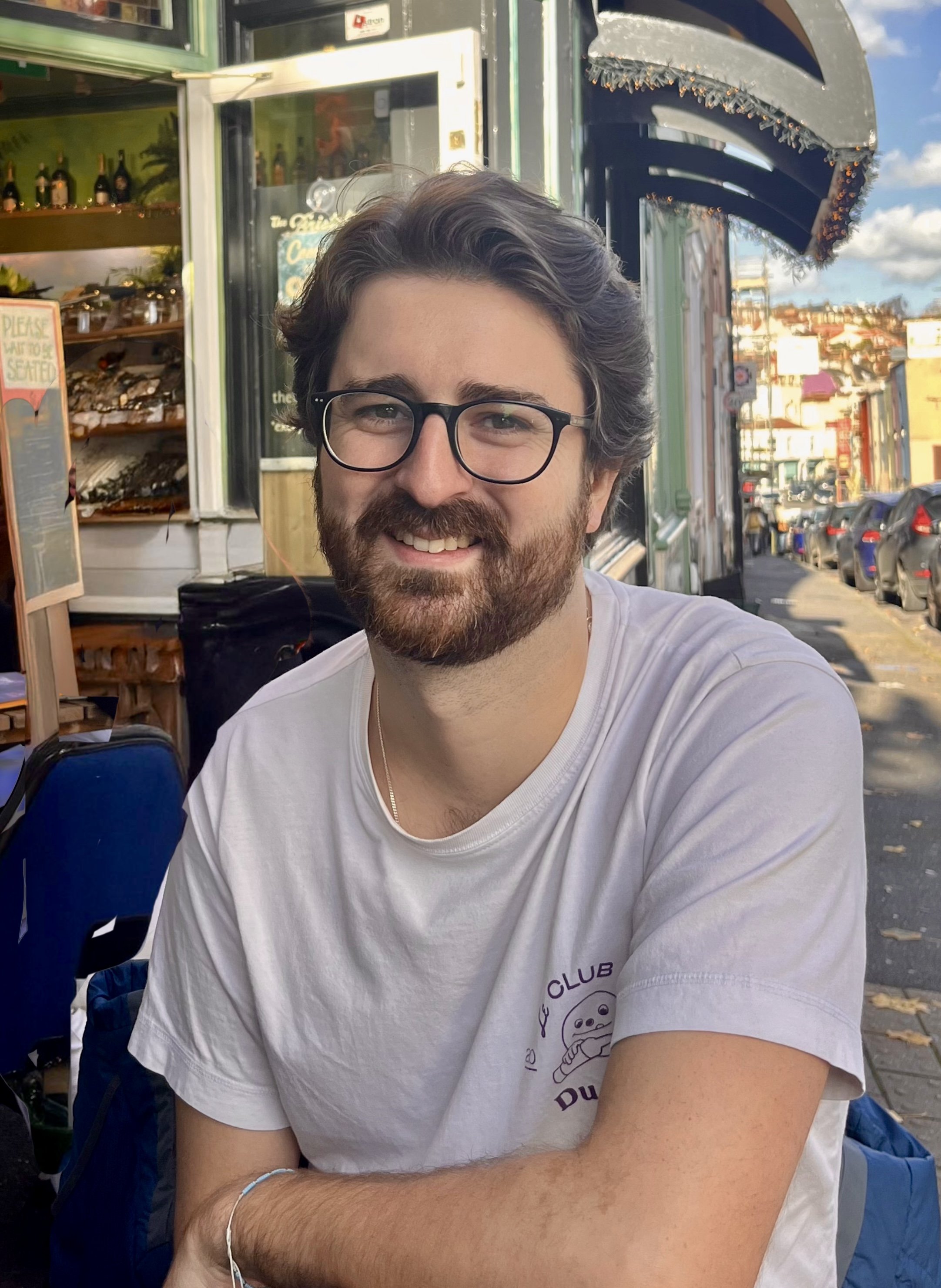 A young man with glasses, a beard, and a white t-shirt sitting outdoors at a cafe or restaurant on a sunny day.