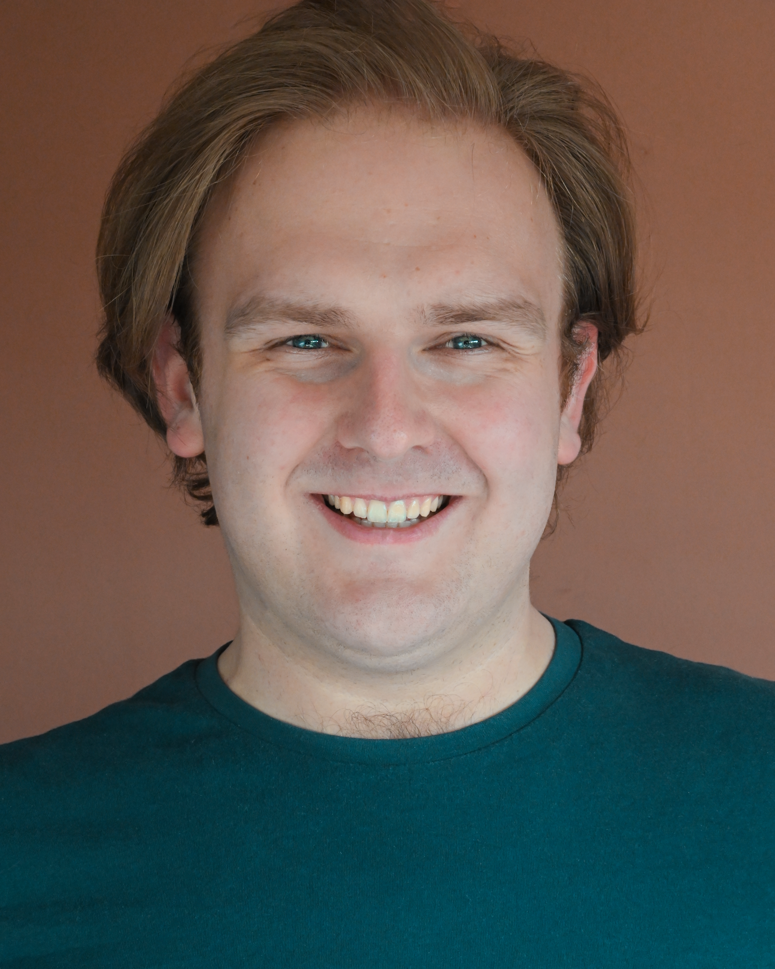 A smiling young man with light skin, light brown hair, and blue eyes, wearing a dark teal shirt against a brown background.